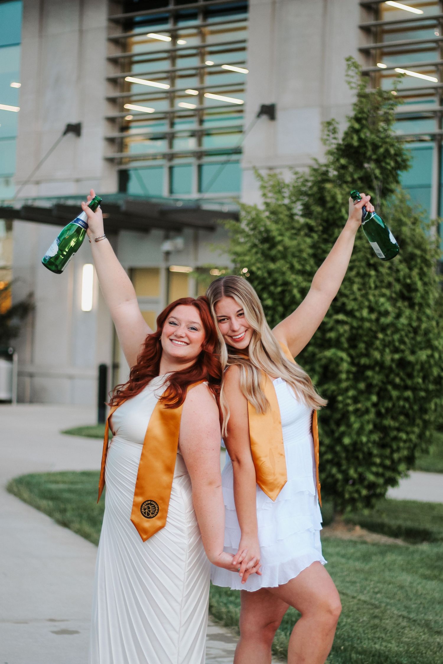 Two Purdue graduates in white dresses and gold stoles celebrate with raised bottles outdoors on Purdue's college campus in West Lafayette