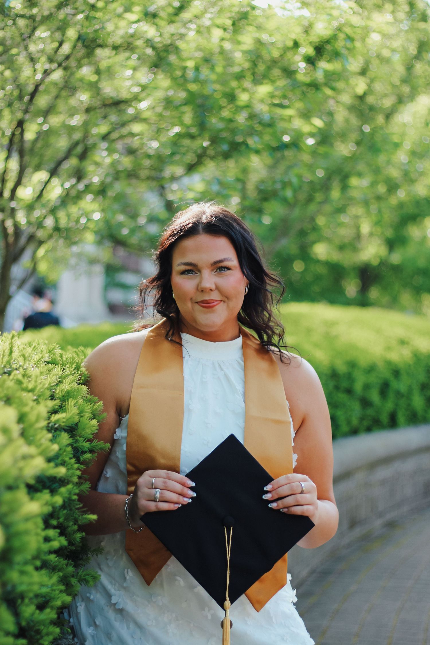 Smiling graduate in white dress holding black cap with gold stole among lush green hedges on Purdue's college campus in West Lafayette