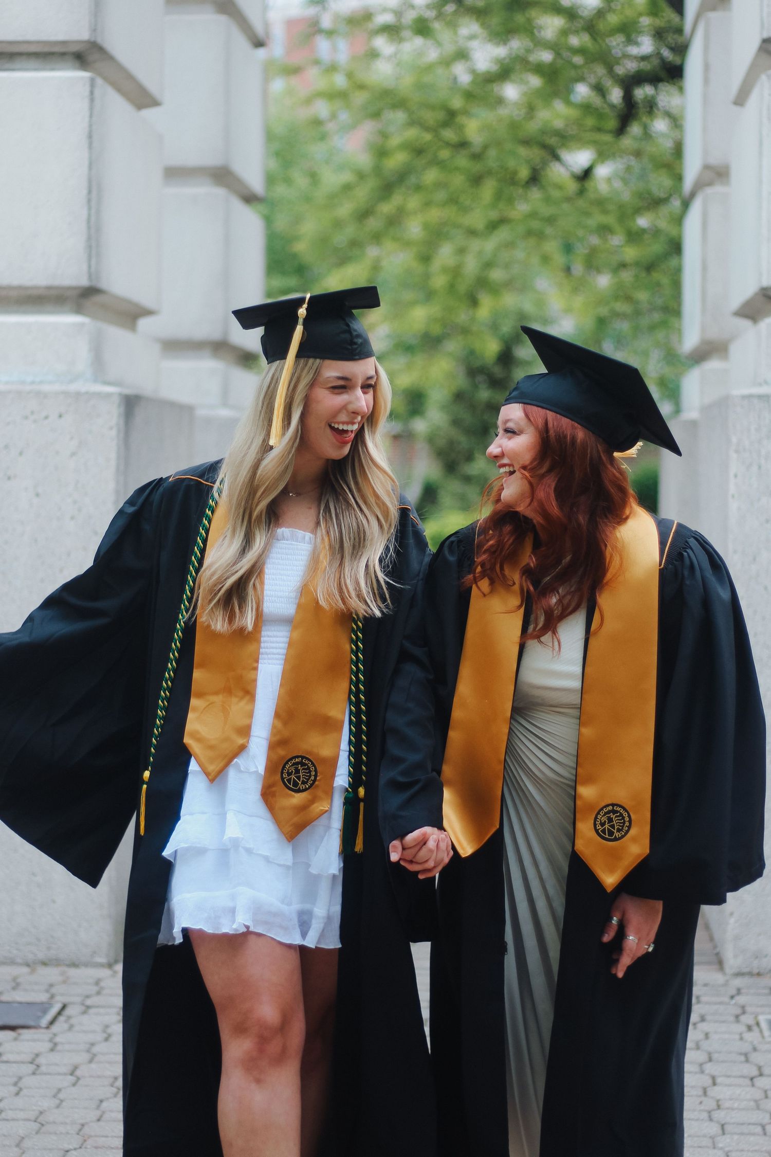 Two smiling graduates in black caps and gowns with gold stoles celebrating together outdoors on Purdue's college campus in West Lafayette