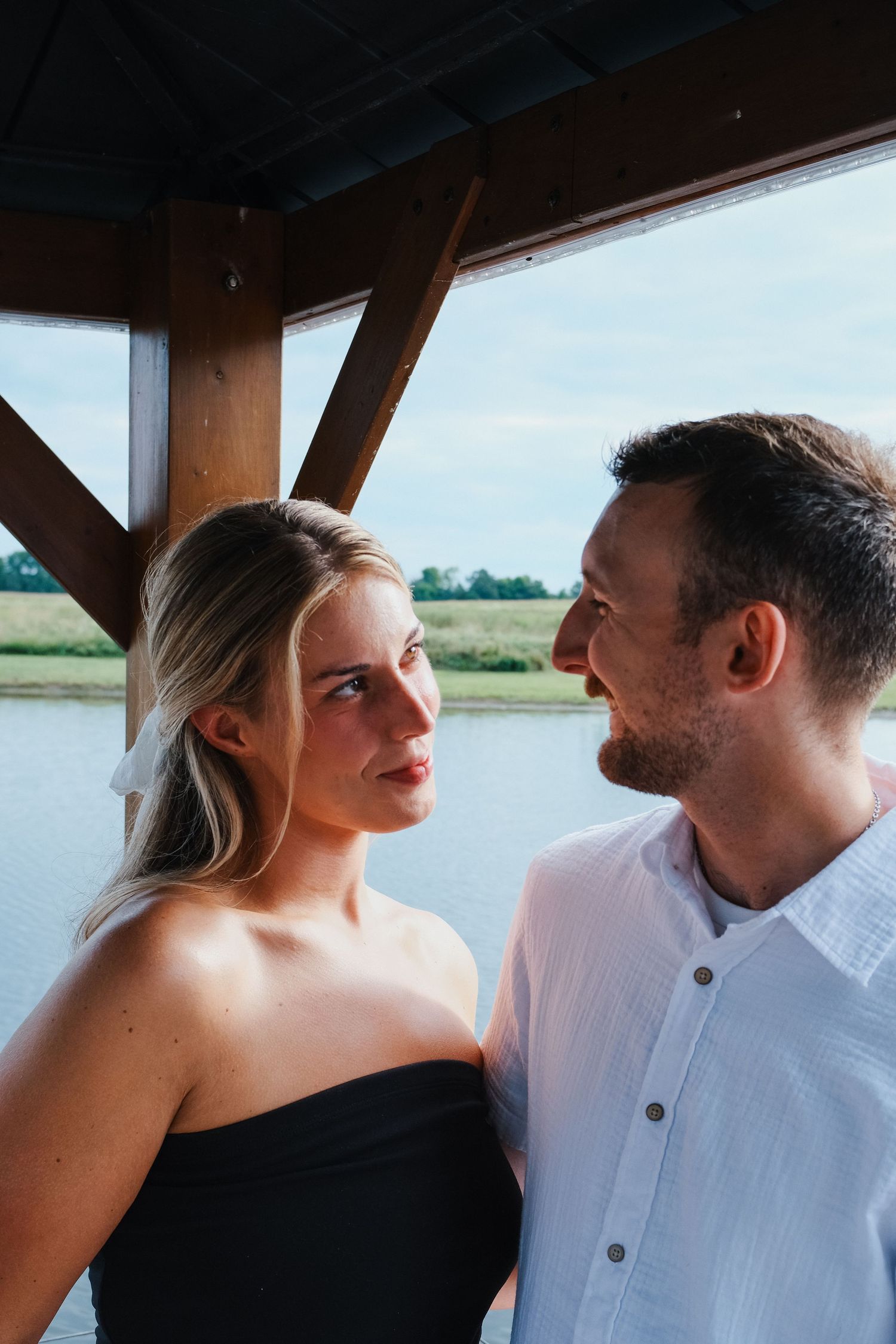A young couple gazes at each other under a wooden gazebo by a lake Indianapolis couple portrait photographer Regan Nichalson Creative