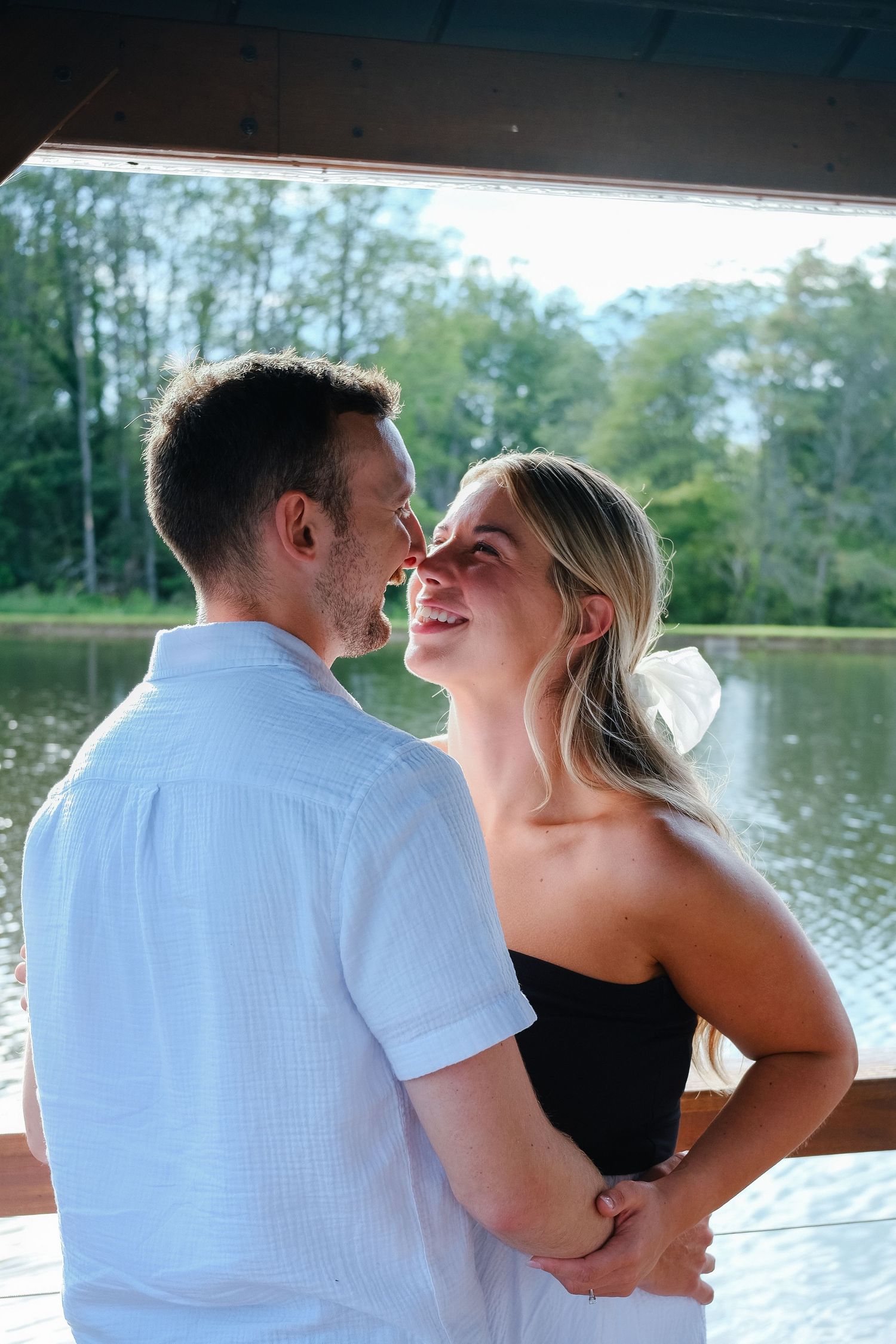 Joyful couple embracing and smiling at each other inside gazebo with sunlit lake backdrop.