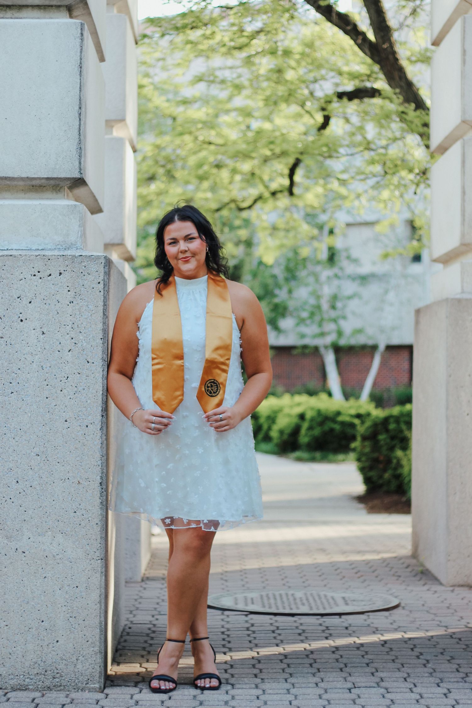 Smiling brunette woman in white dress with gold graduation stole posing between pillars outdoors on Purdue's college campus