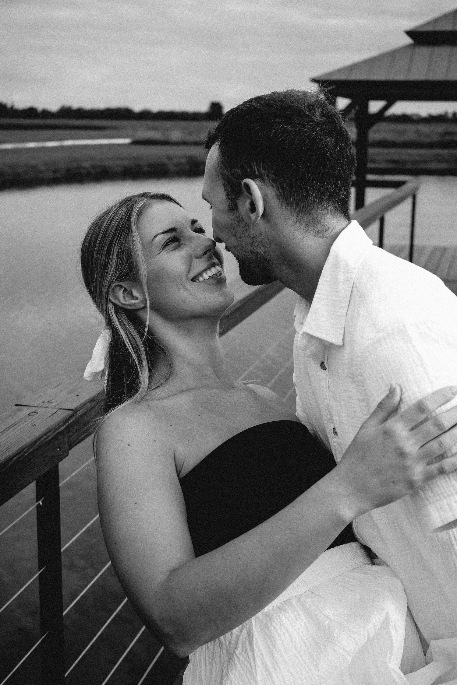 Black and white photo of a smiling woman in a black strapless dress being kissed by a man on a dock.