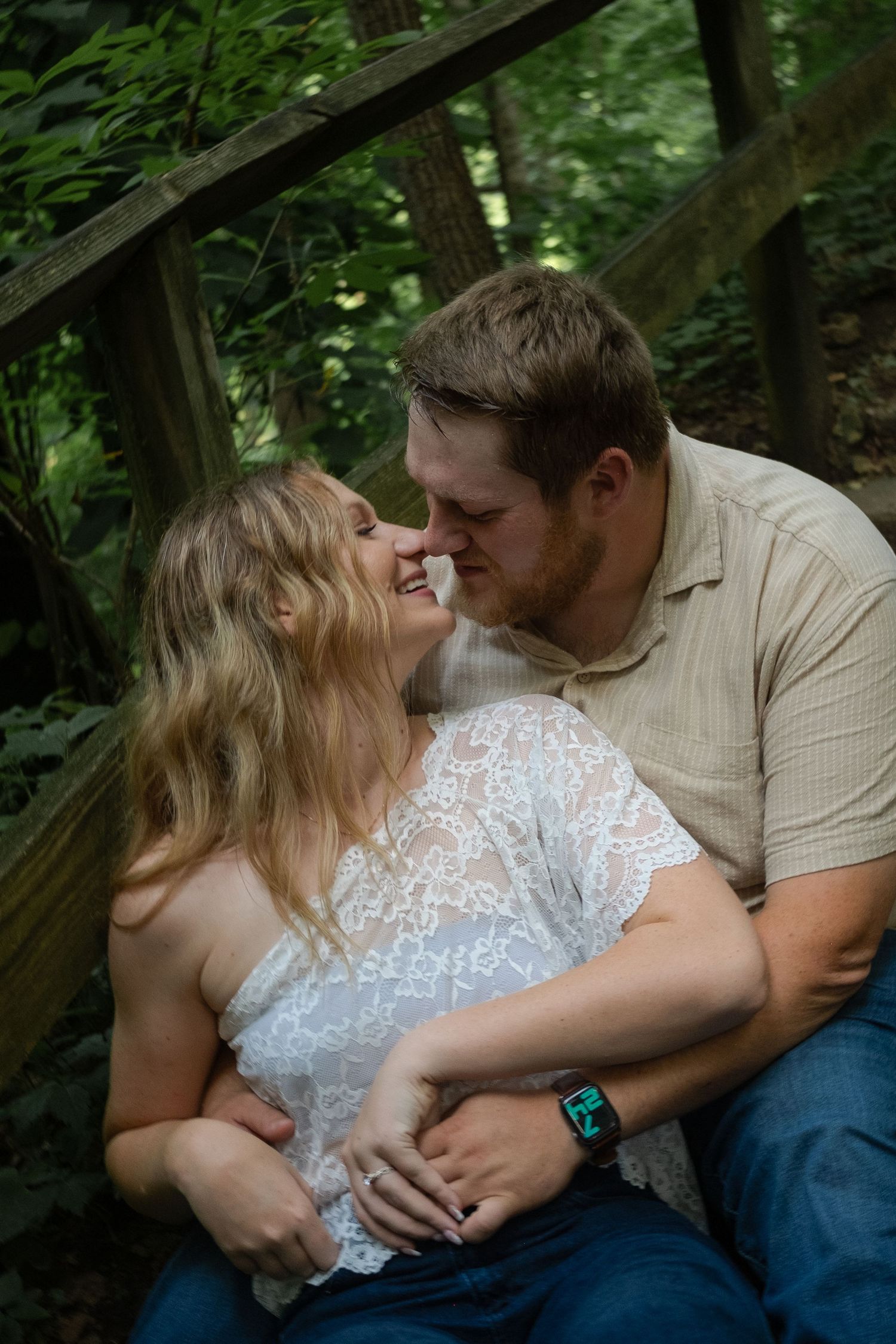 A couple shares a romantic moment outdoors on wooden steps surrounded by lush green trees.