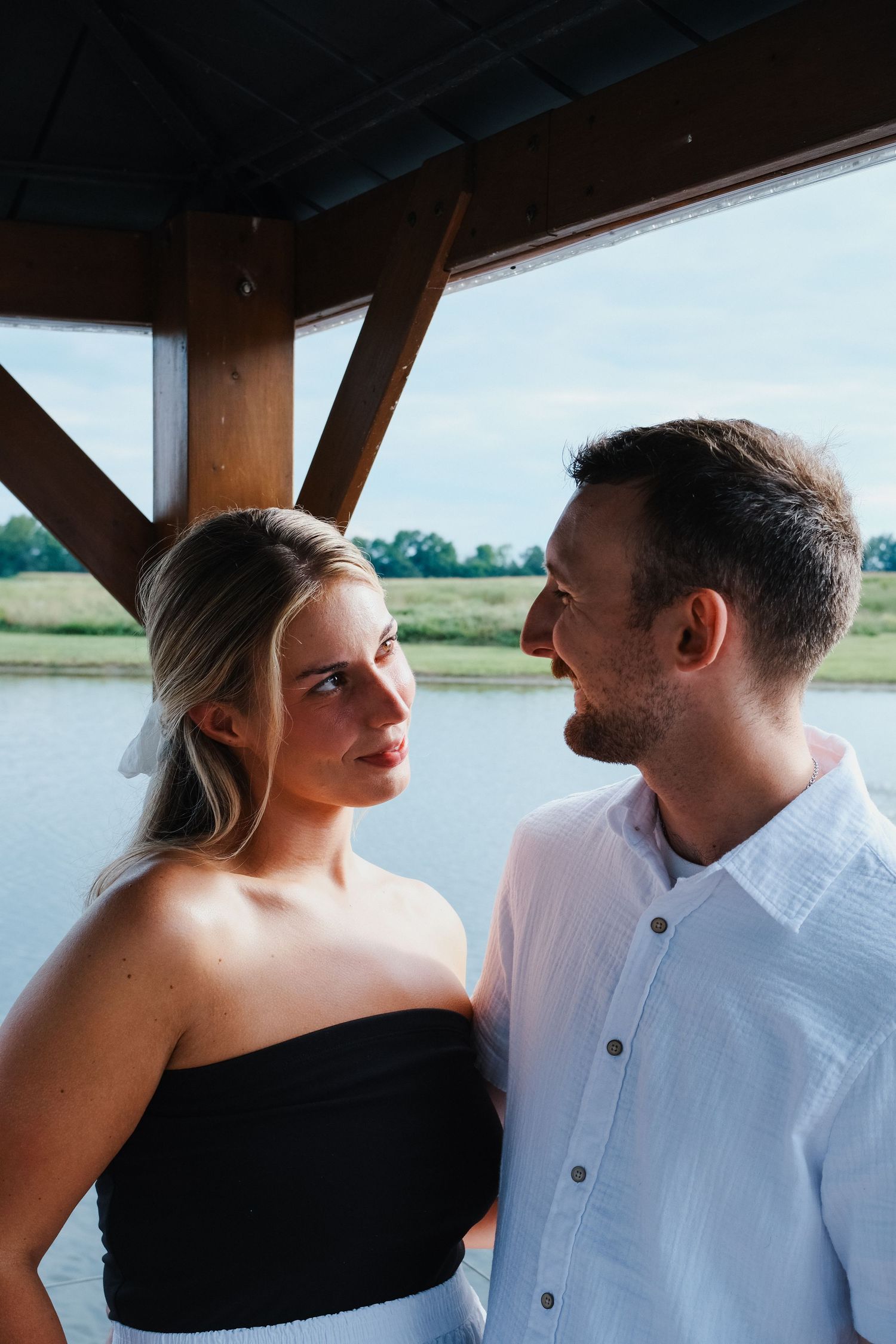 Couple in black and white outfits gazing at each other under wooden structure by lake.