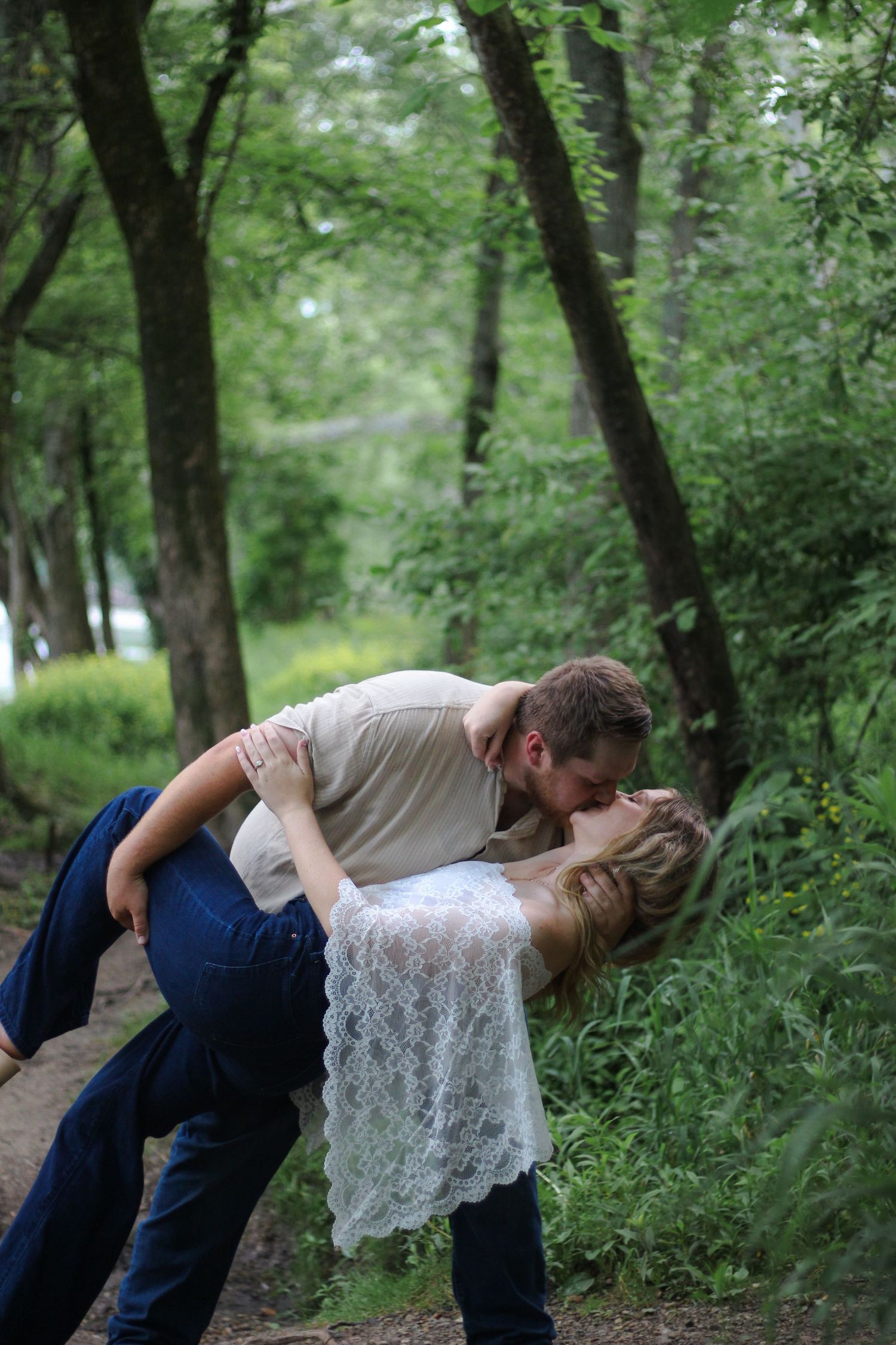 A couple shares a romantic dip kiss on a forest path surrounded by lush green trees.
