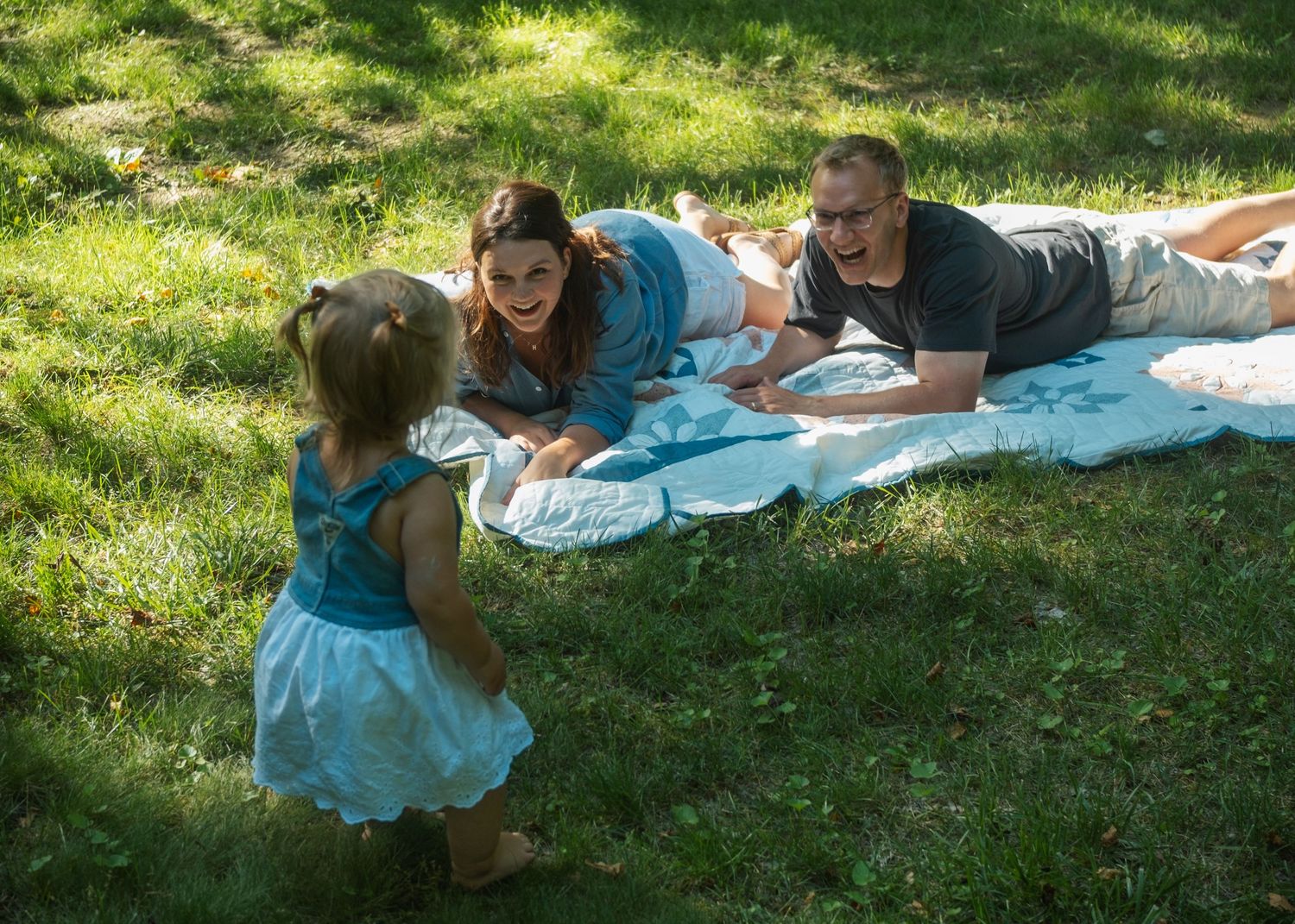 Family enjoying a sunny outdoor picnic on a blanket in the grass with a young girl in a blue dress.