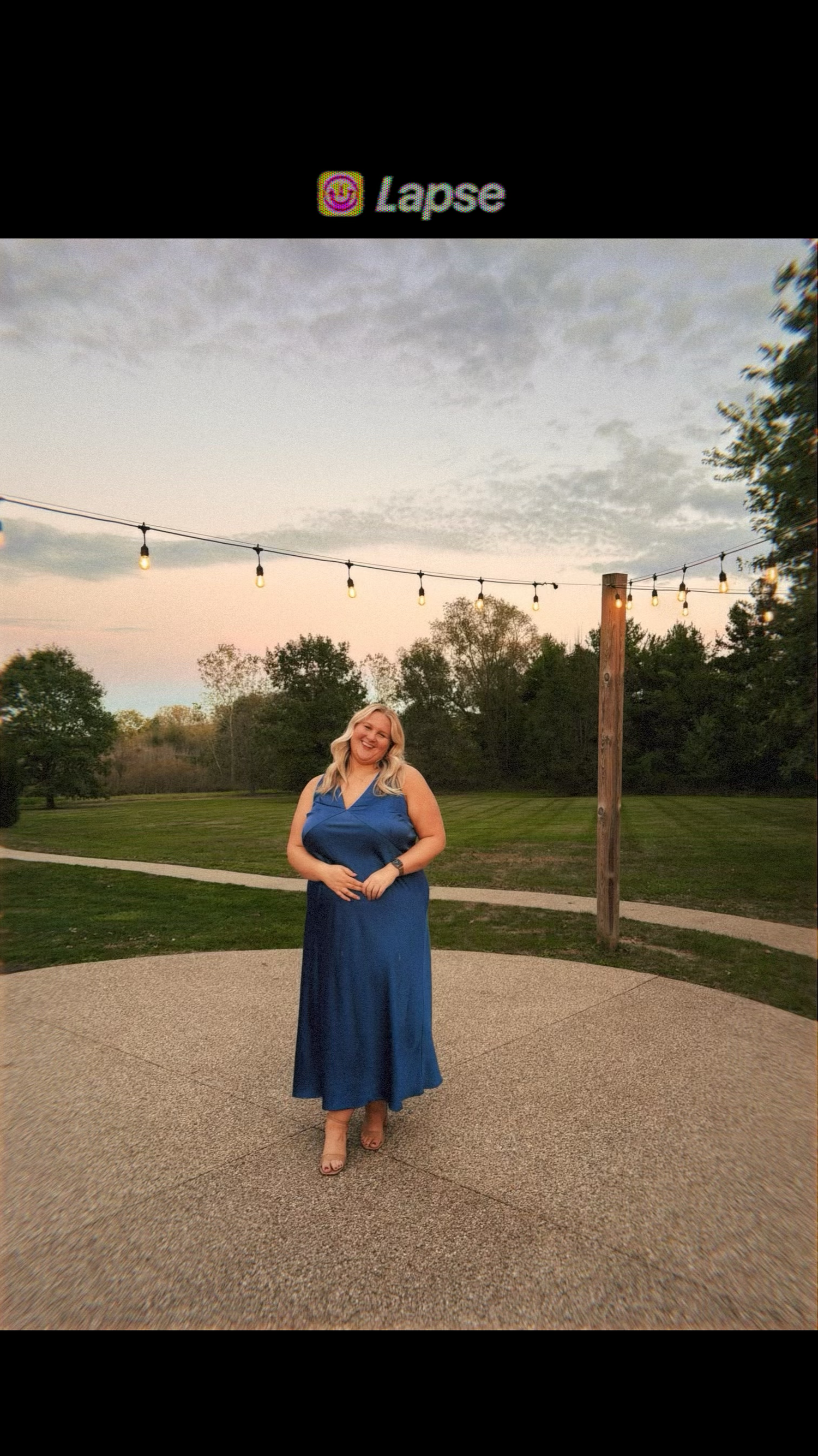 Blonde woman in elegant blue maxi dress posing under string lights at an outdoor venue at sunset. Carmel family photographer Regan Nichalson
