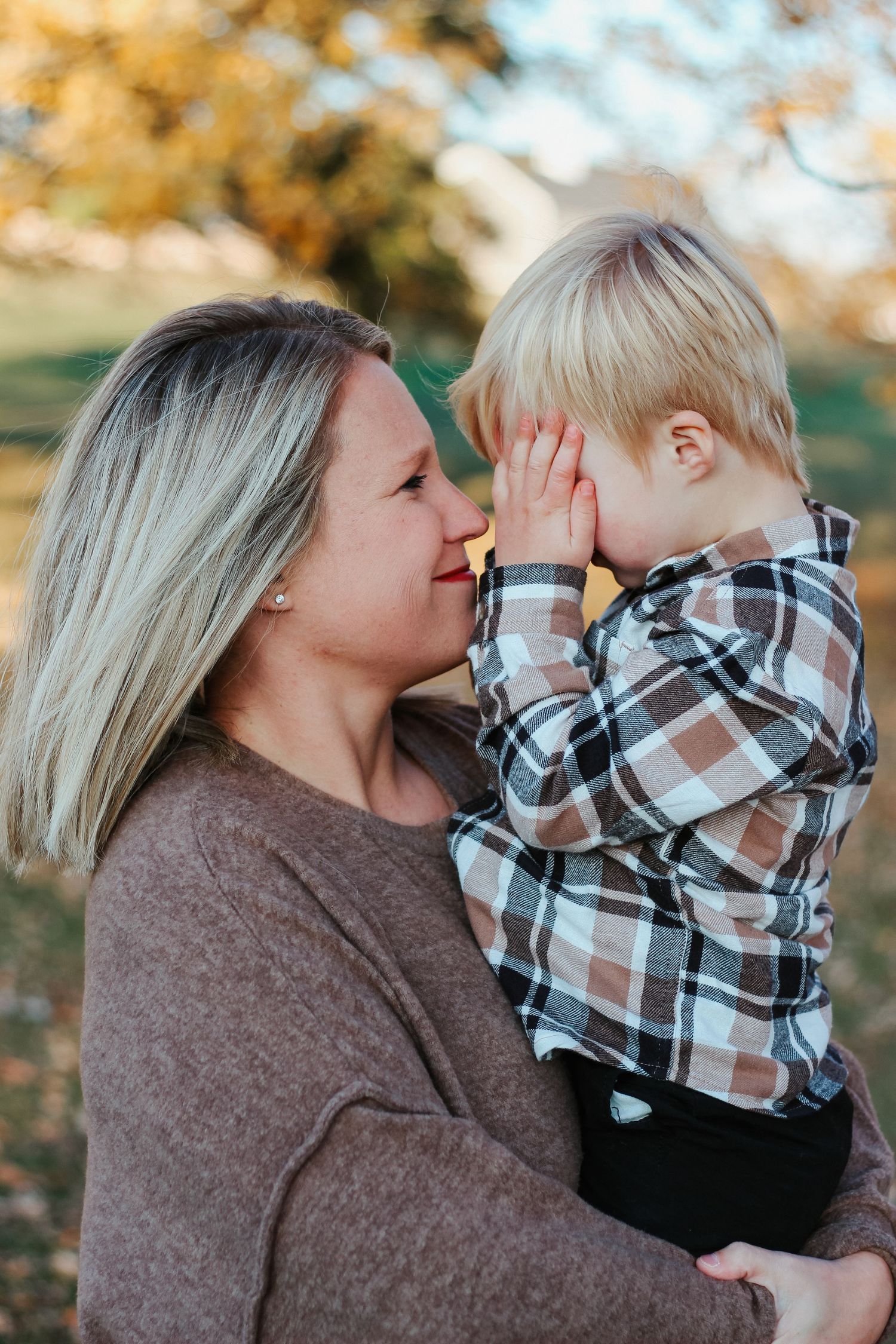 Loving mother holding young blonde toddler boy in plaid shirt in autumn park setting.