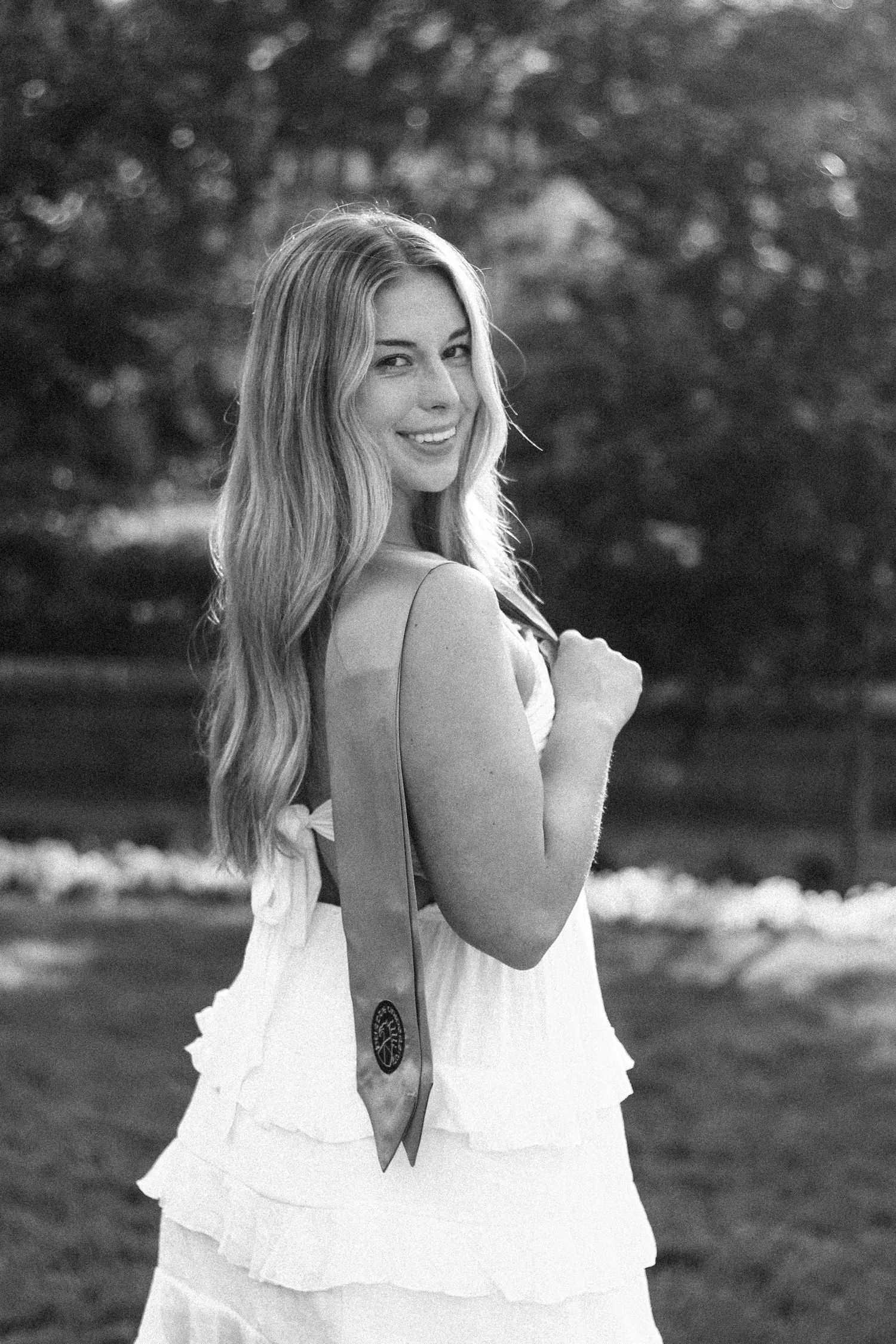 Black and white photo of smiling blonde woman in white ruffled dress holding graduation stole on Purdue's college campus in West Lafayette