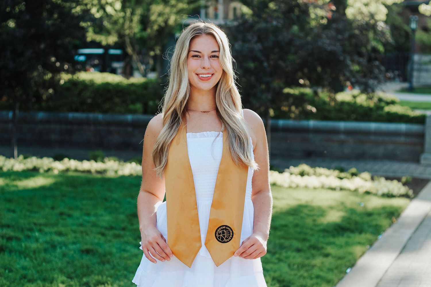 Smiling blonde woman in white dress wearing gold graduation stole standing on sunny campus grounds on Purdue's college campus in West Lafaye