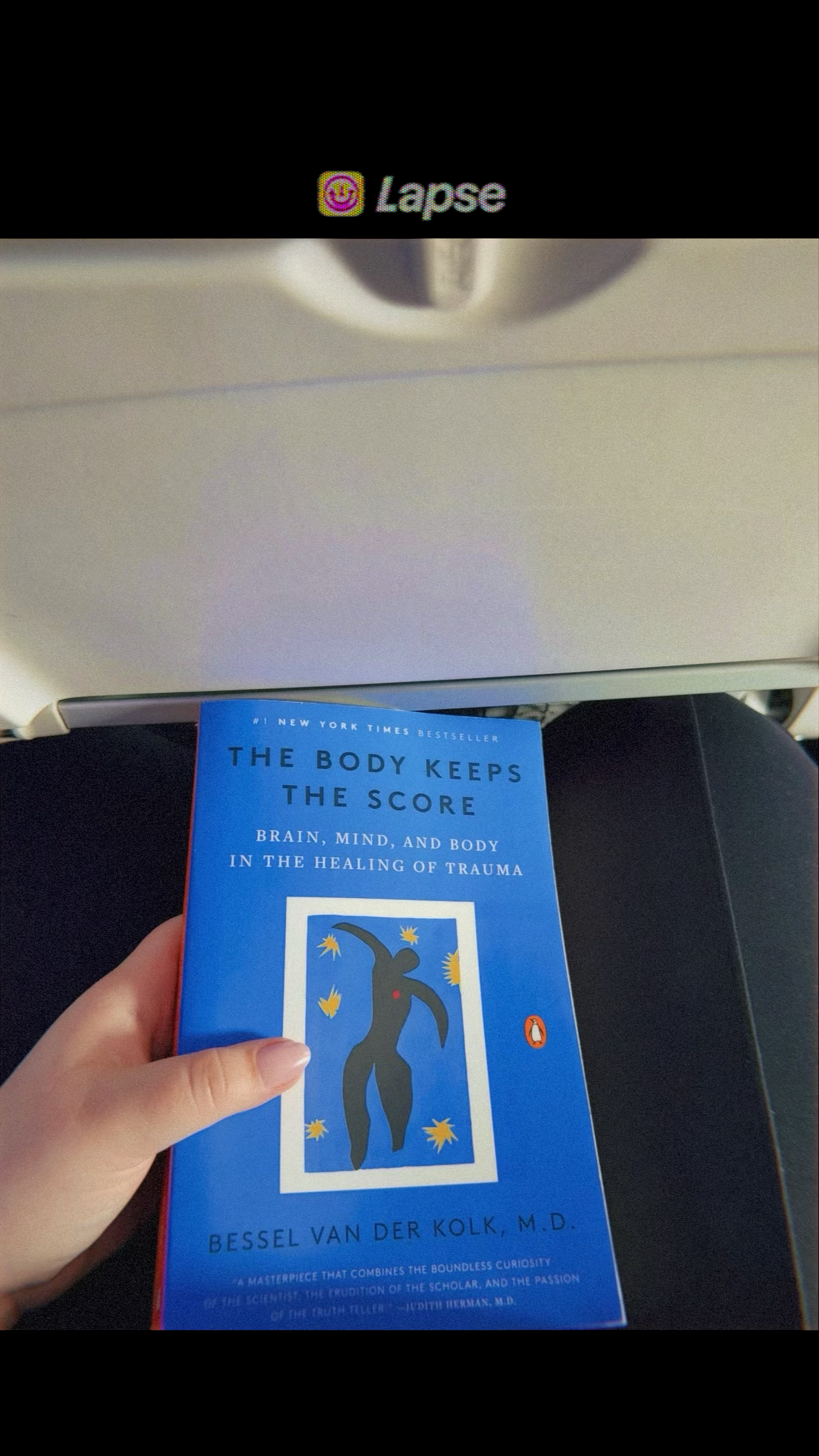 Hand holding the book The Body Keeps the Score by Bessel van der Kolk on an airplane tray table. Carmel family photographer Regan Nichalson