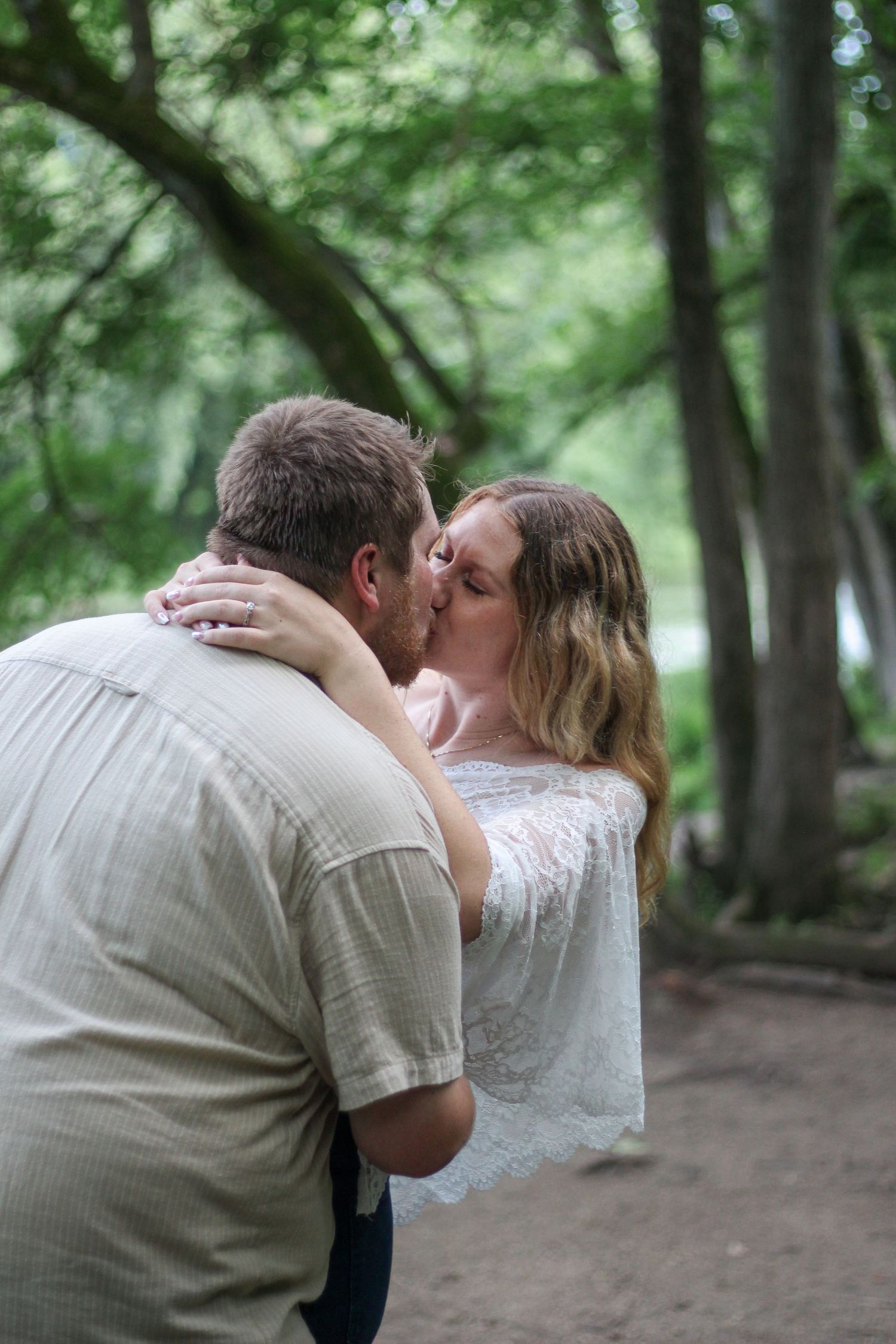 Couple sharing a romantic kiss outdoors in a lush green forest setting, woman in white lace top embracing man in beige shirt.