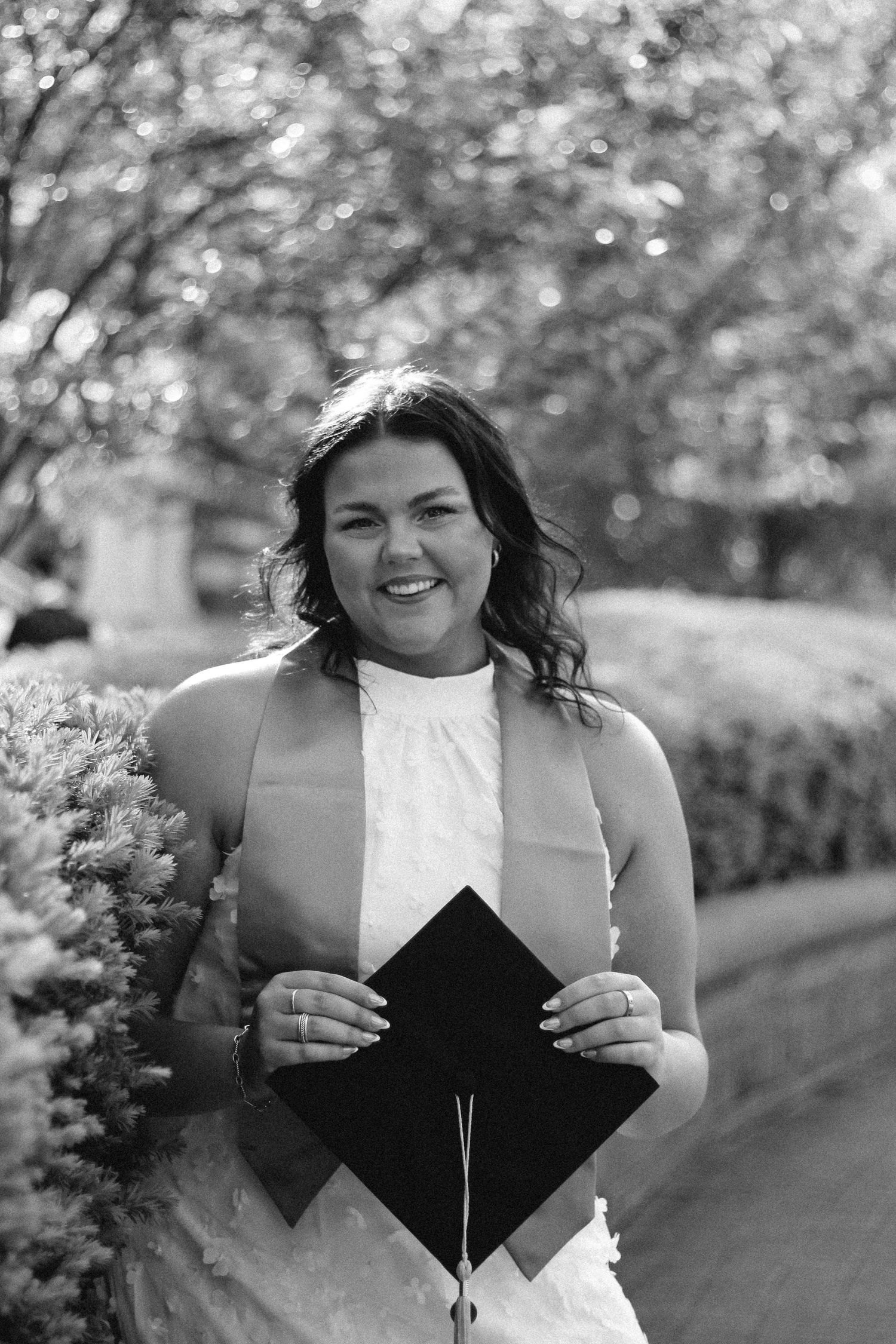 Black and white photo of smiling brunette woman holding graduation cap outdoors. on Purdue's college campus in West Lafayette