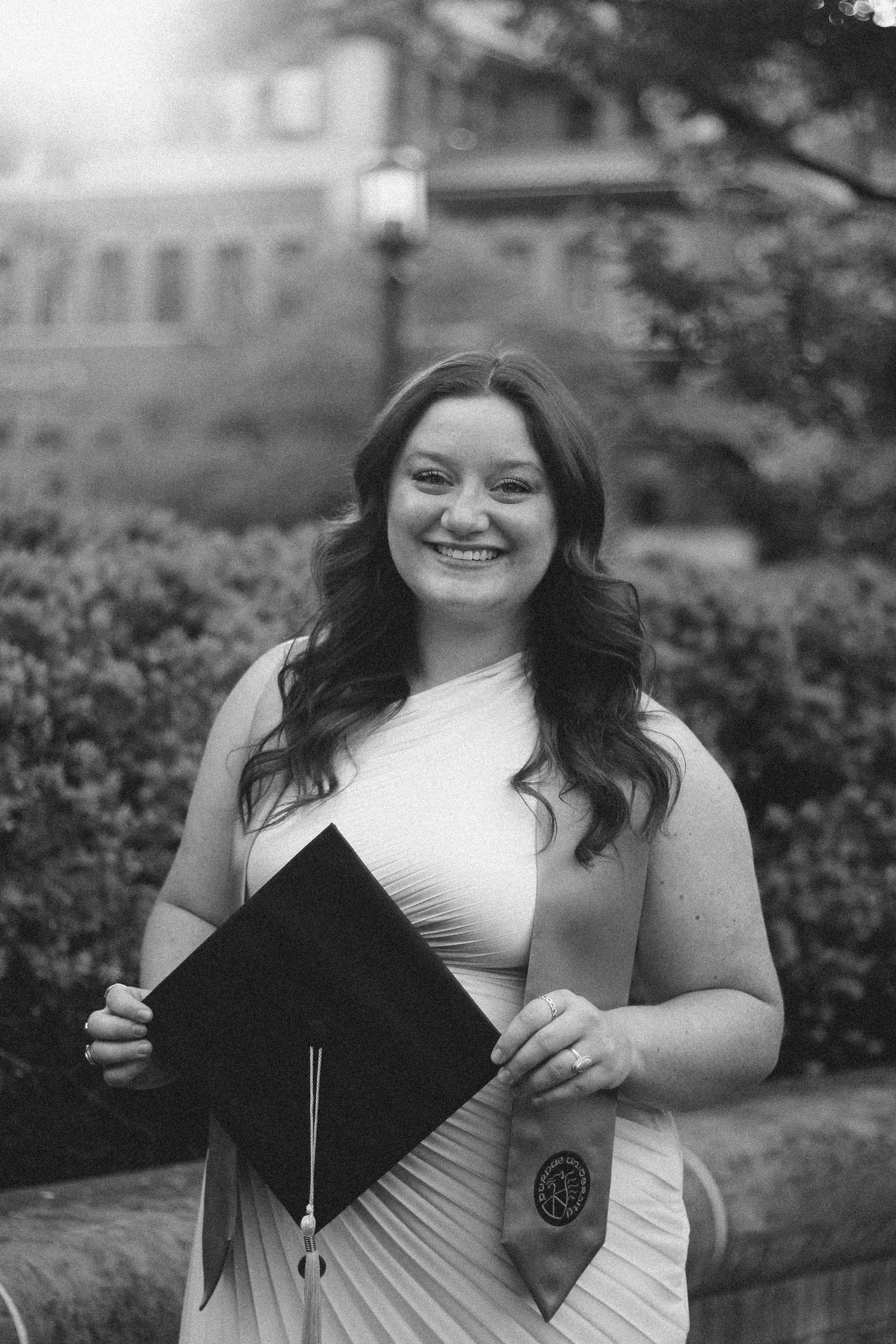 Black and white photo of smiling woman in white dress holding graduation cap with stole on Purdue's college campus in West Lafayette