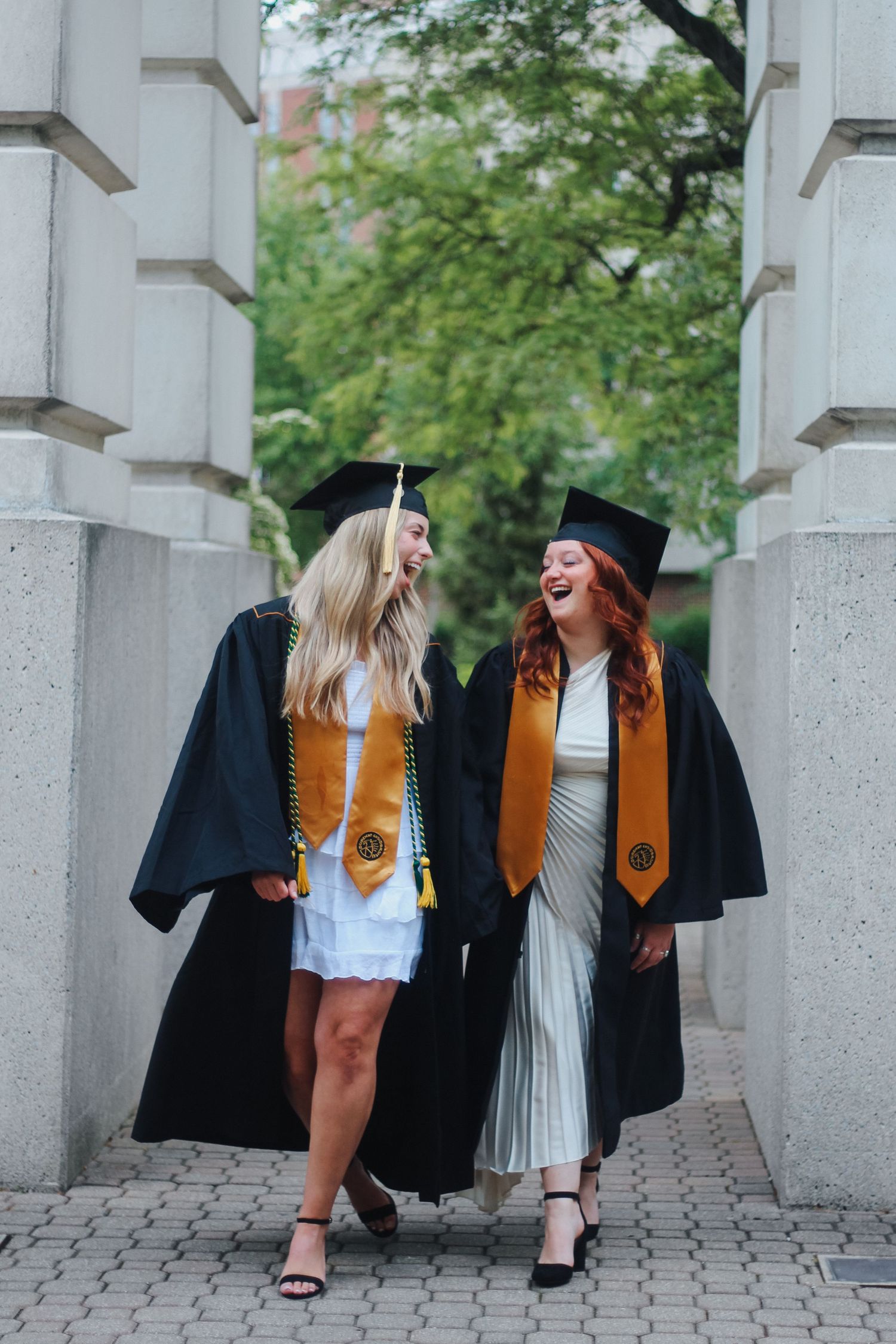 Two laughing graduates in black caps and gowns with gold stoles walking together on campus on Purdue's college campus in West Lafayette