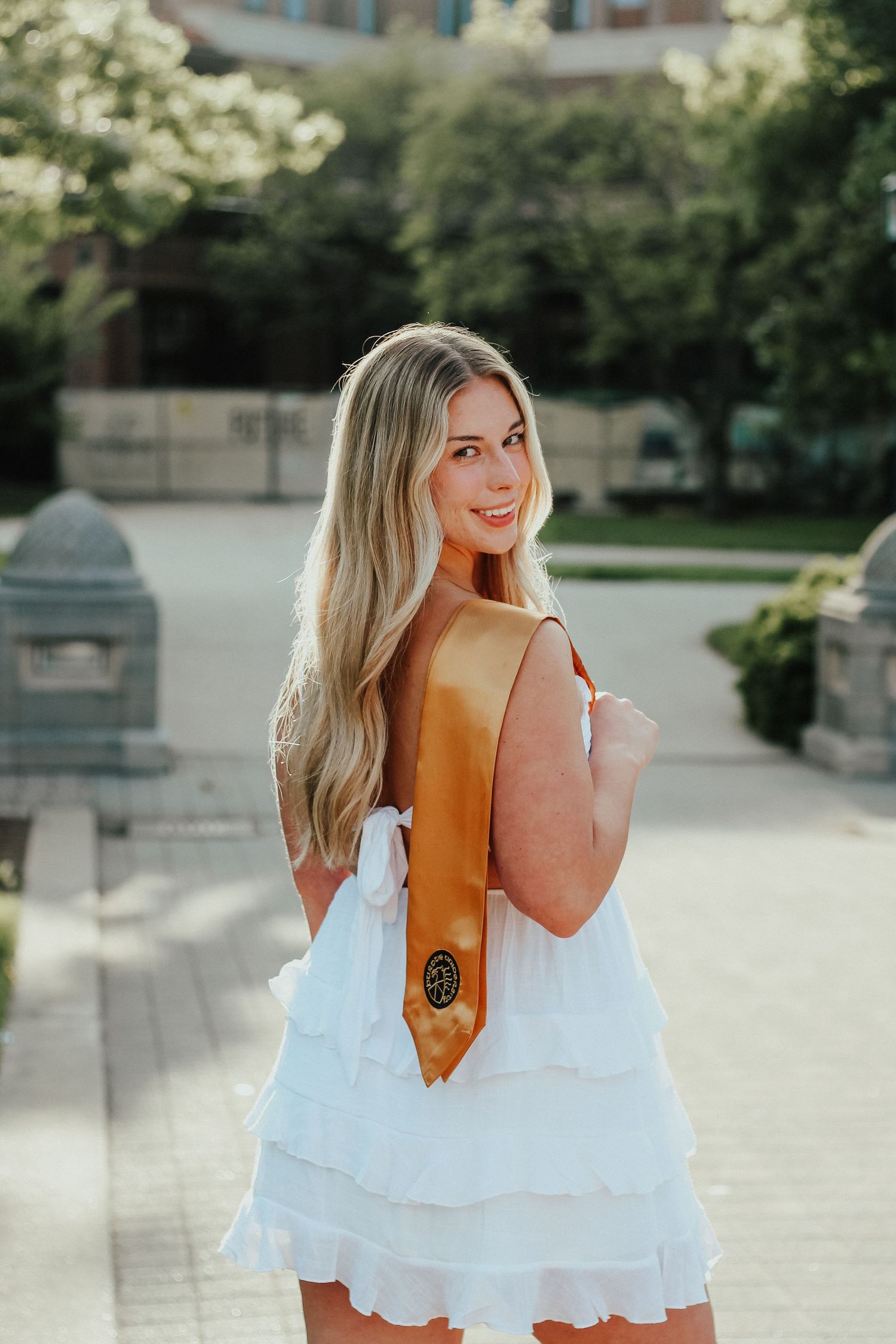 Smiling blonde woman in white dress holding gold graduation stole outdoors on campuson Purdue's college campus in West Lafayette