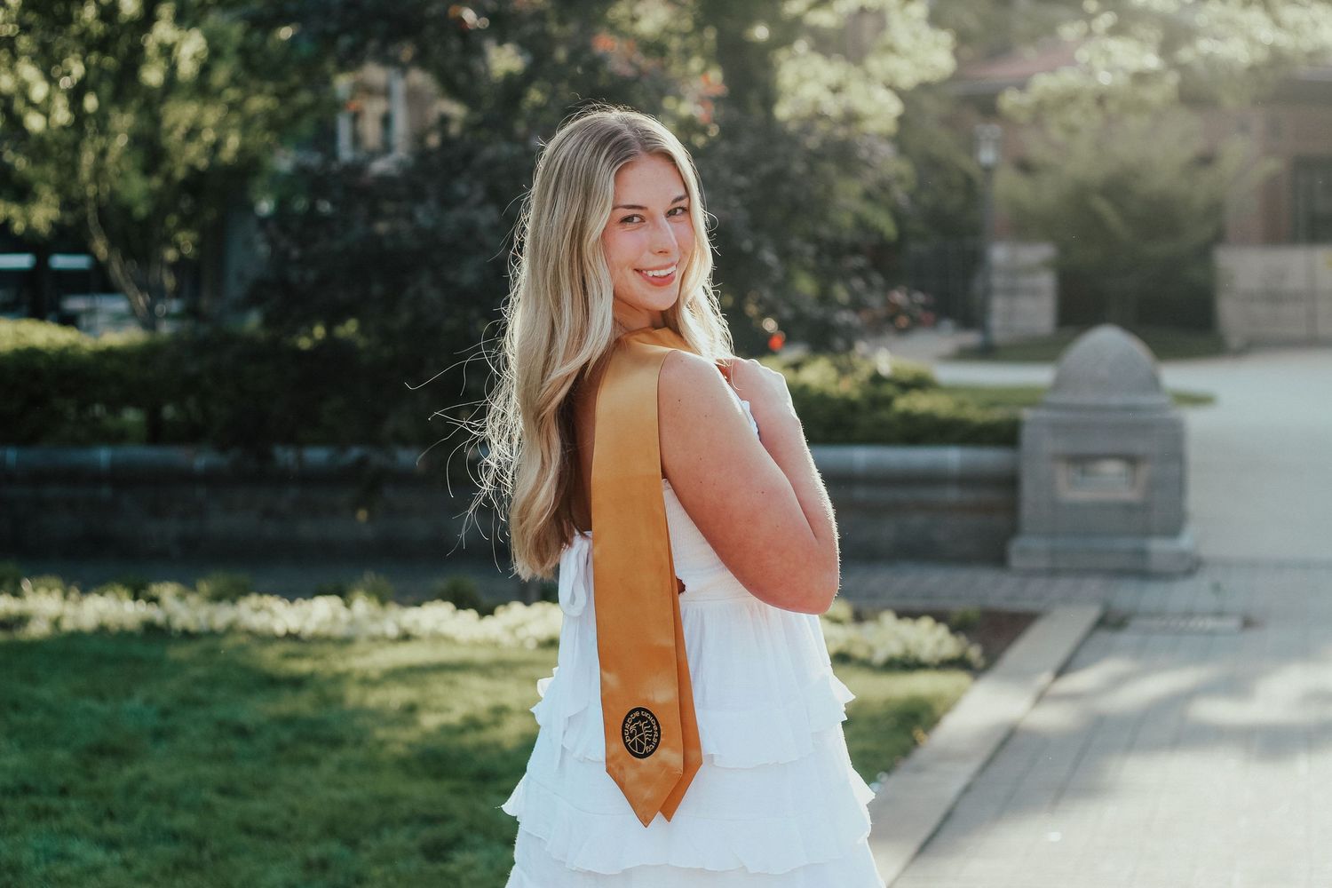 Smiling blonde woman in white dress with gold graduation stole posing outdoors on Purdue's college campus in West Lafayette