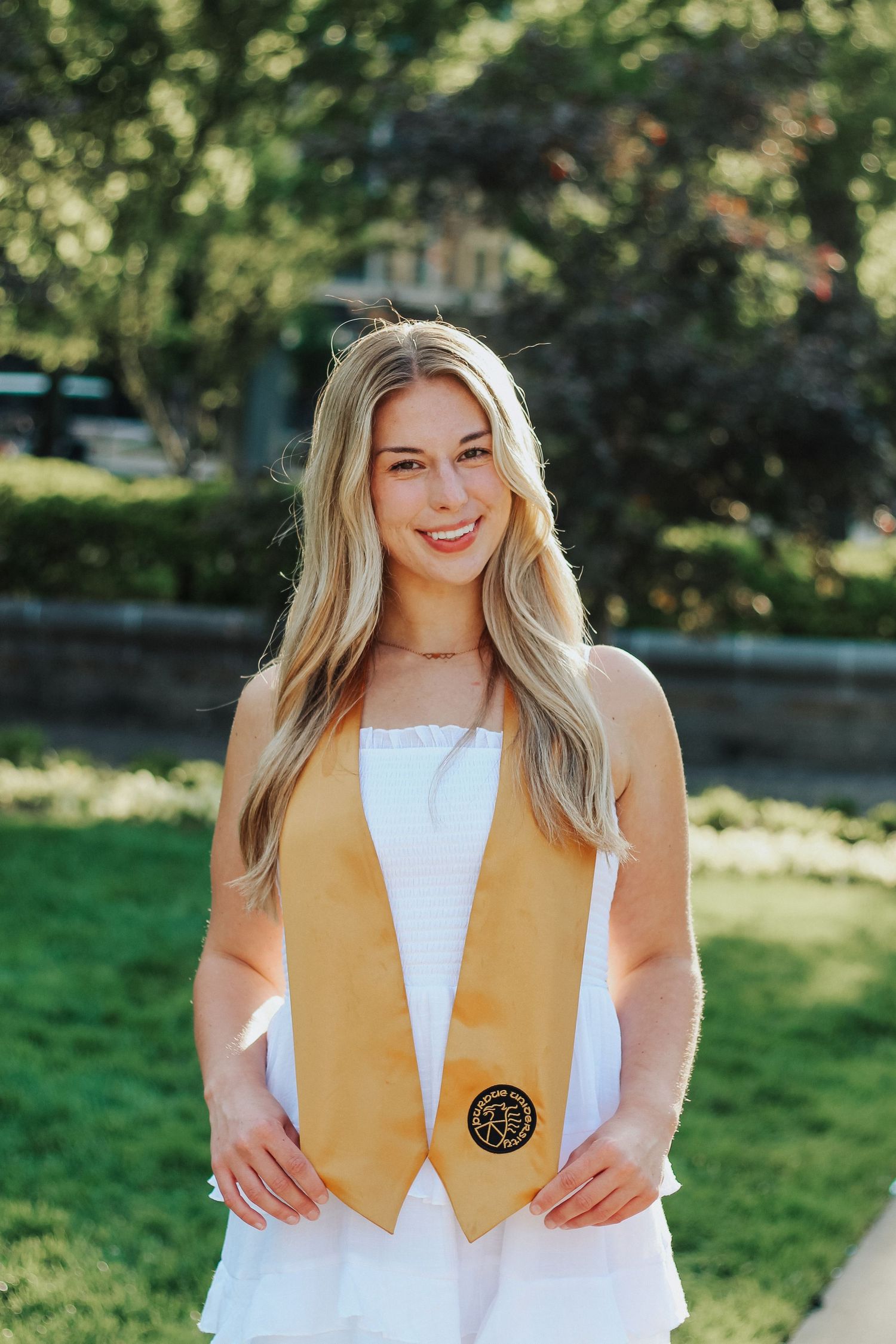 Smiling blonde woman in white dress wearing gold graduation stole standing on sunny campus lawn on Purdue's college campus in West Lafayette