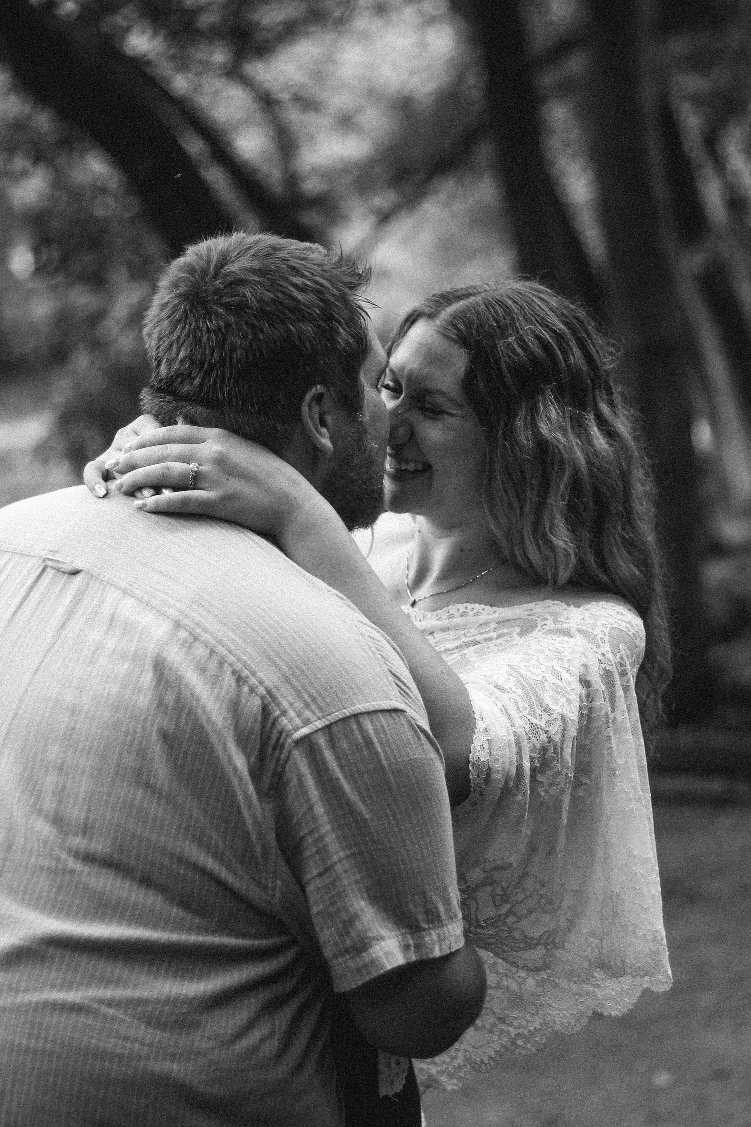 Black and white photo of a smiling woman embracing a man outdoors among trees, wearing a lace top and ring.