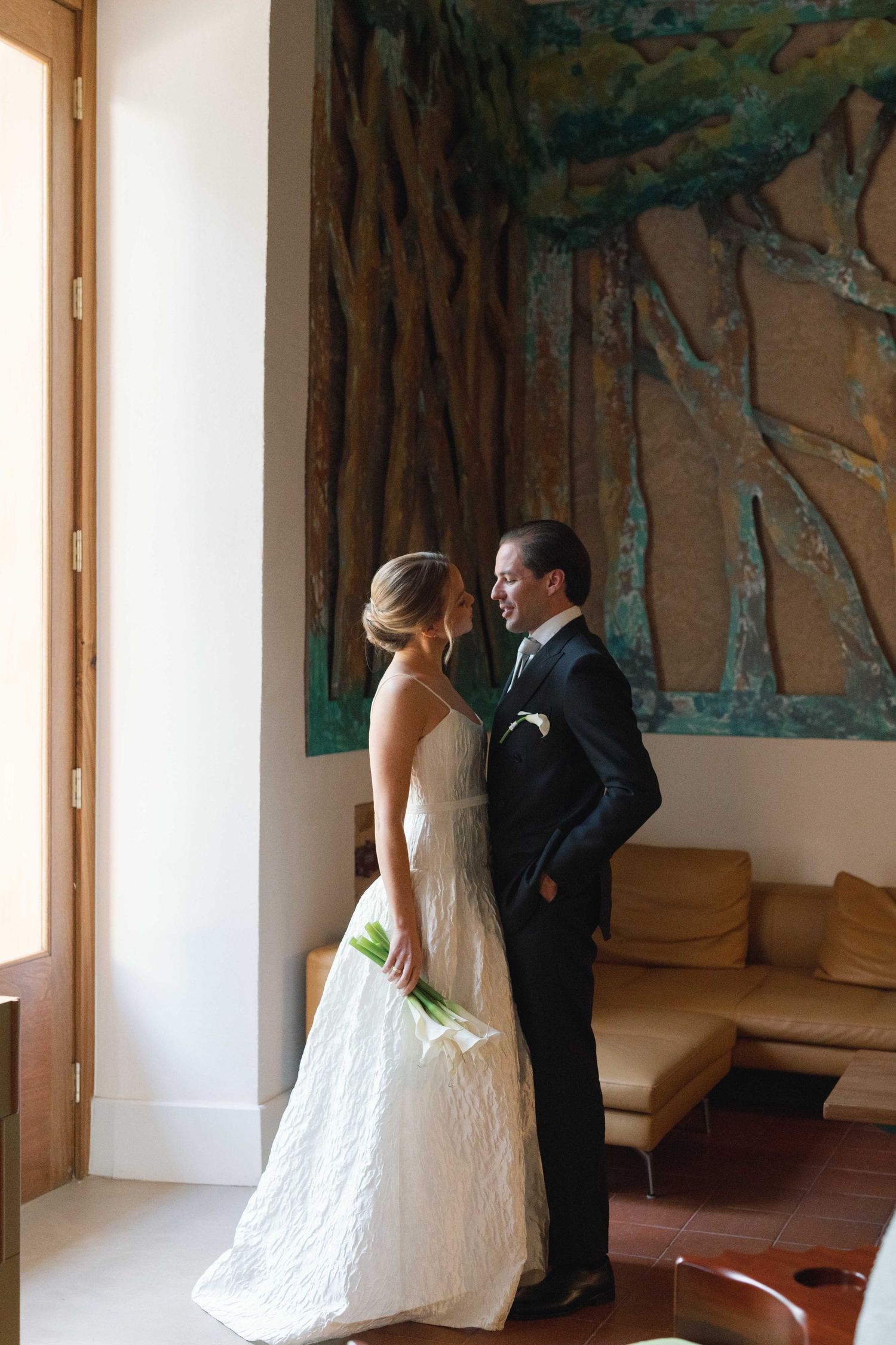 The bride and groom framed by natural light in a refined interior. A calm, intimate moment defined by understated luxury.