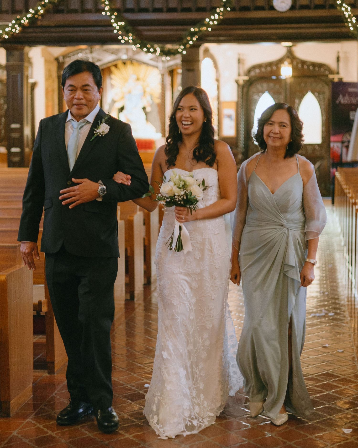 Wedding party walks down decorated church aisle with string lights overhead.