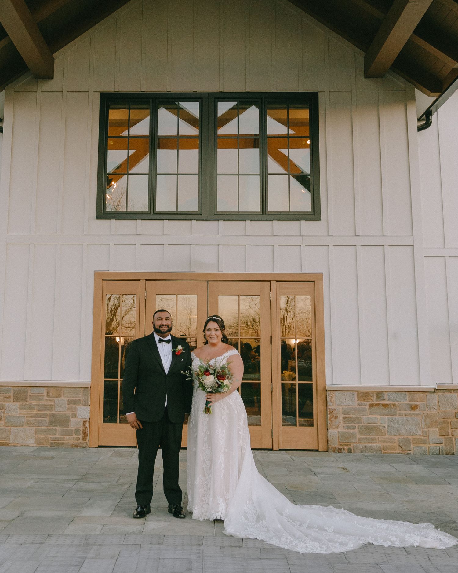 A wedding couple poses in front of a modern white barn-style building with architectural details.