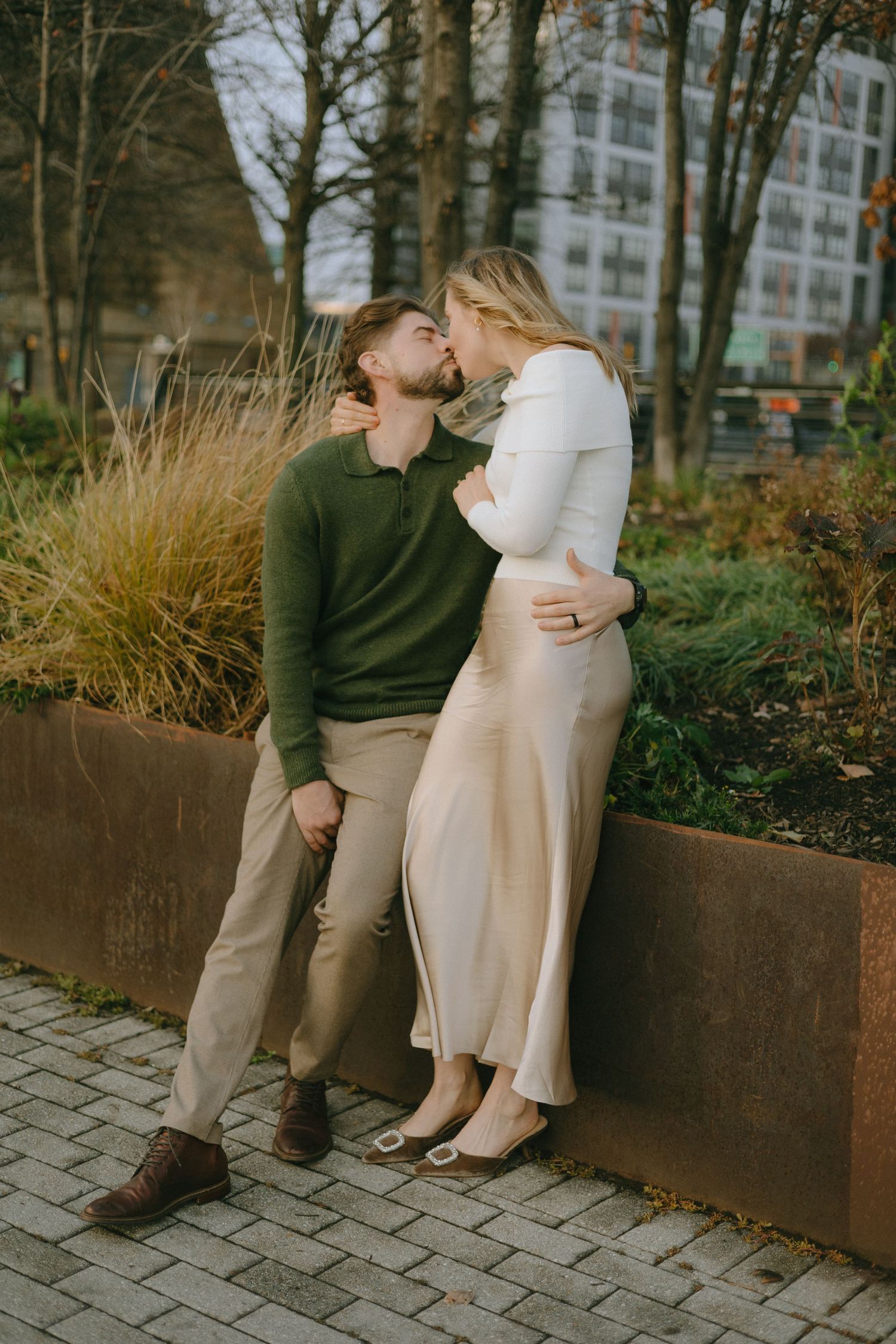 A couple sits together on a rustic metal bench surrounded by autumn foliage and stone pavers.