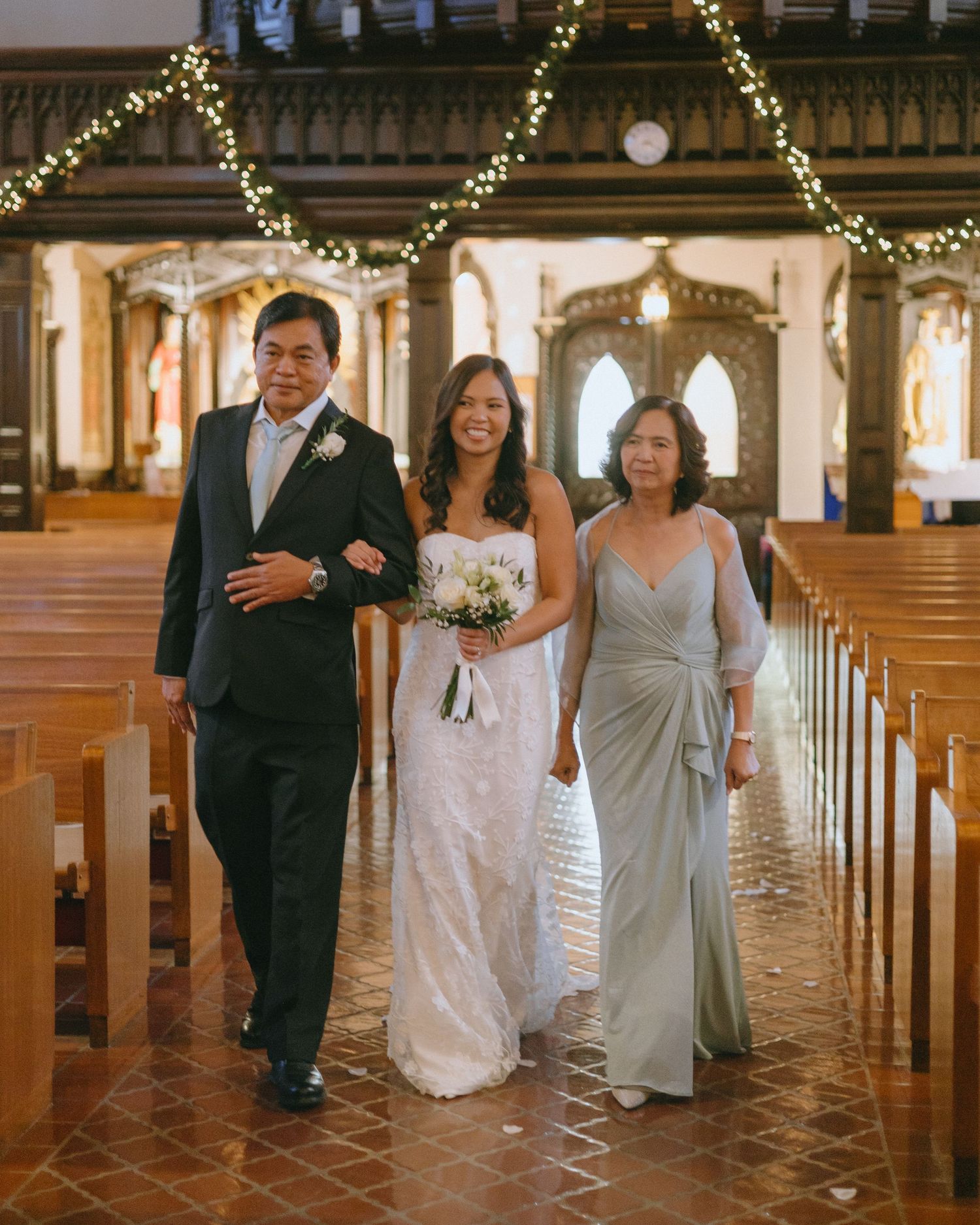 Bridal party proceeds down church aisle with festive Christmas lights overhead.