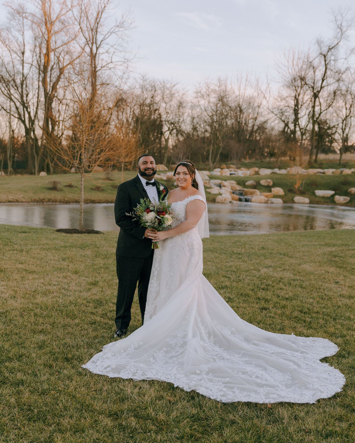 Newlyweds stand together by a pond with a stunning lace wedding dress train.
