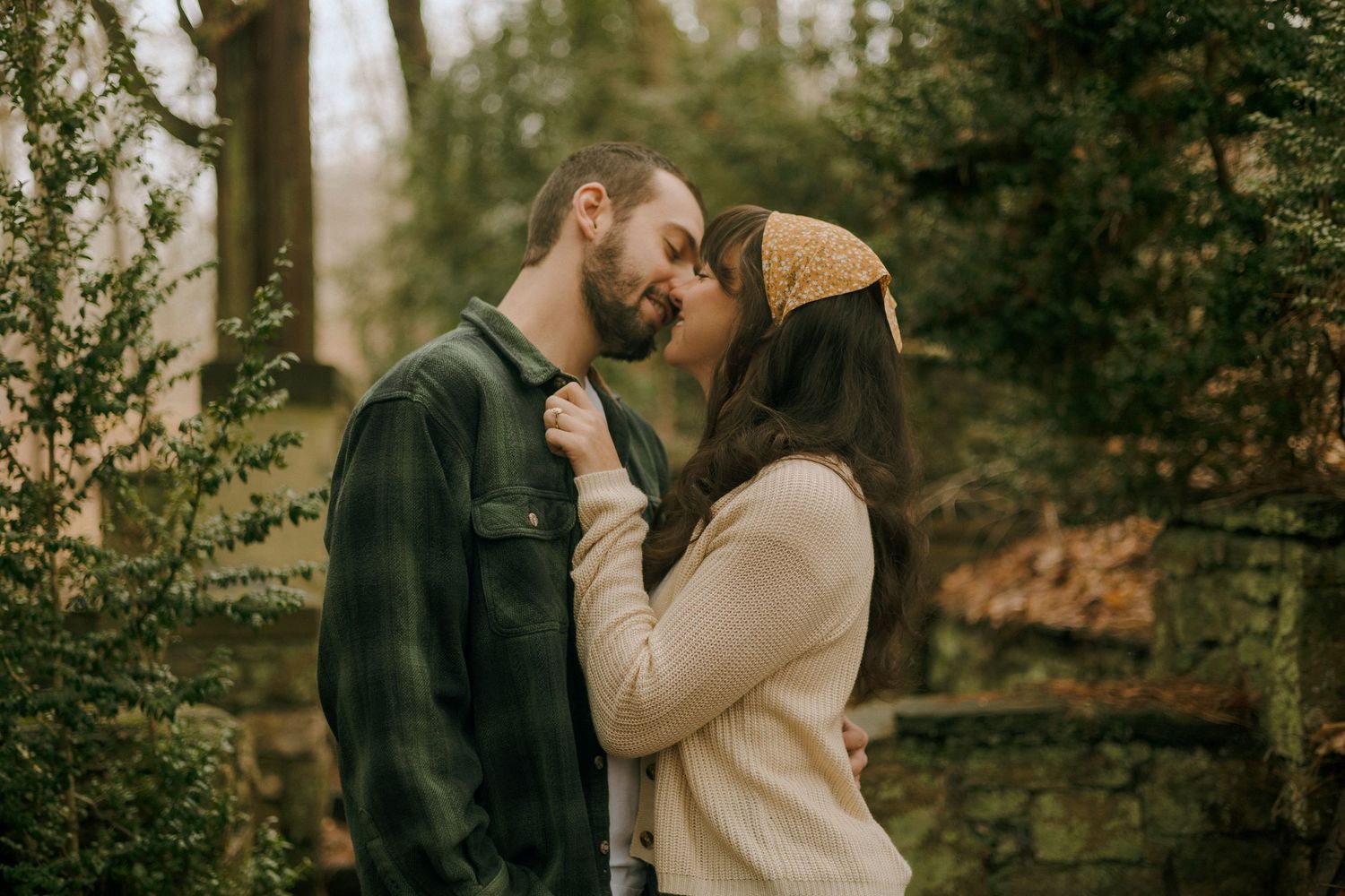 A romantic couple shares an intimate moment in a wooded setting during autumn with stone walls in the background.