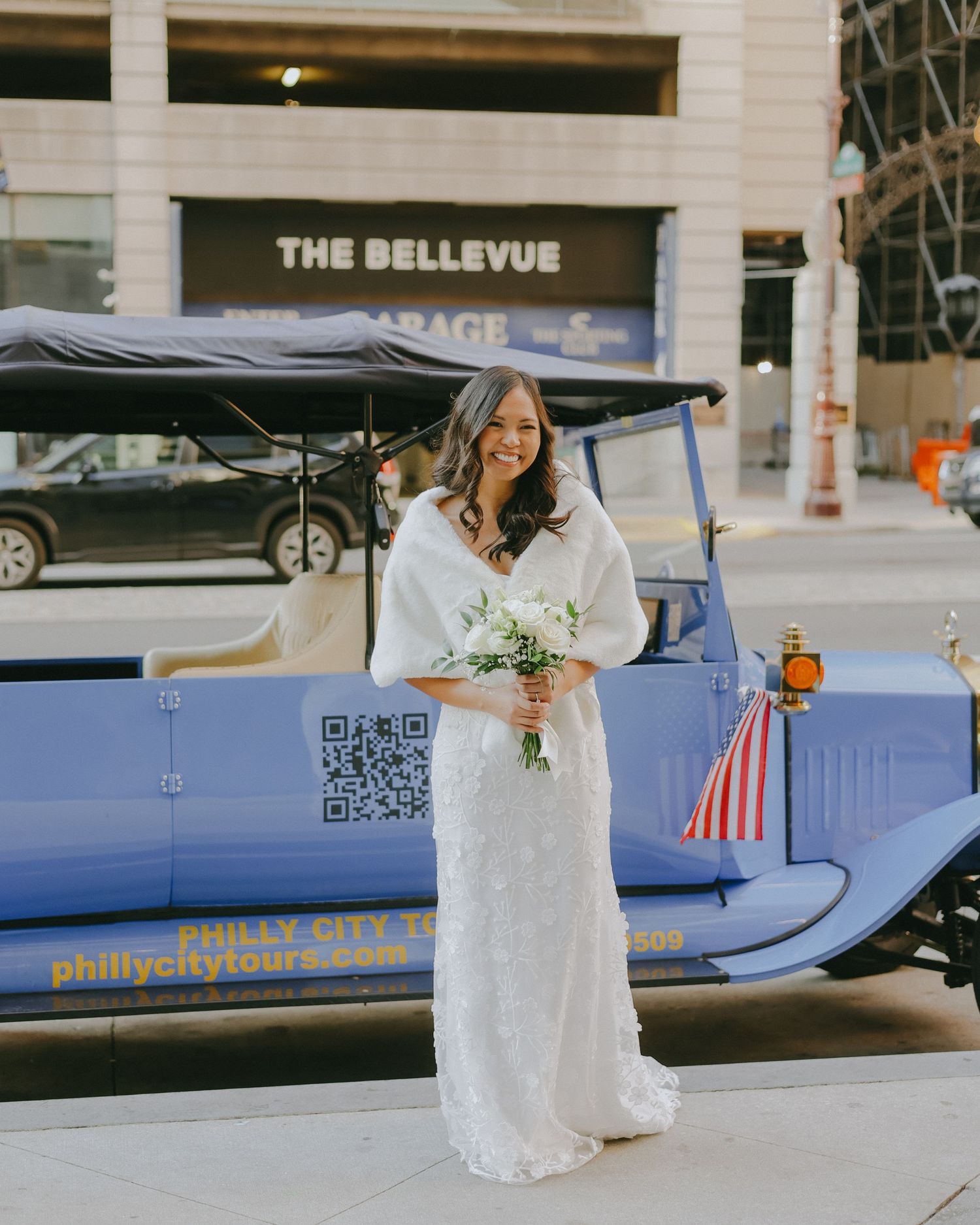 A bride in a lace wedding dress sits on a blue bench outside The Bellevue building.