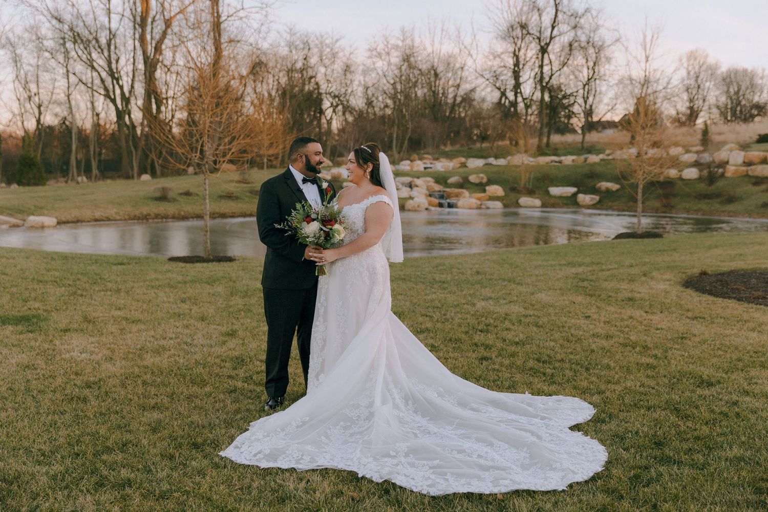 A couple poses outdoors in formal wedding attire near a scenic lake during golden hour.