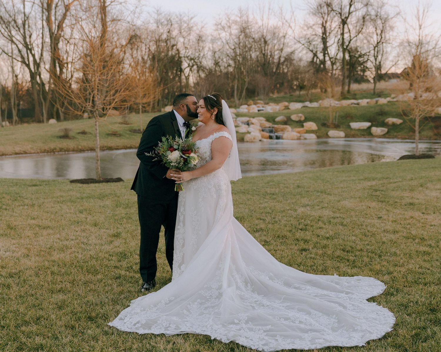 A wedding couple poses by a scenic pond with fall trees in the background.
