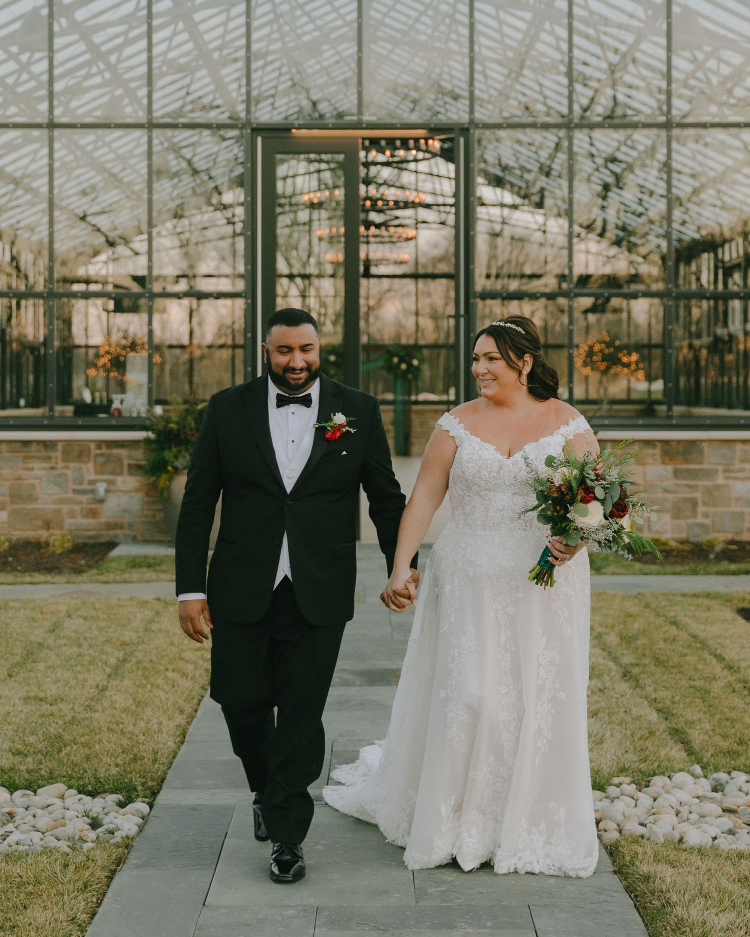 Newlyweds hold hands while walking along a path with a greenhouse backdrop.