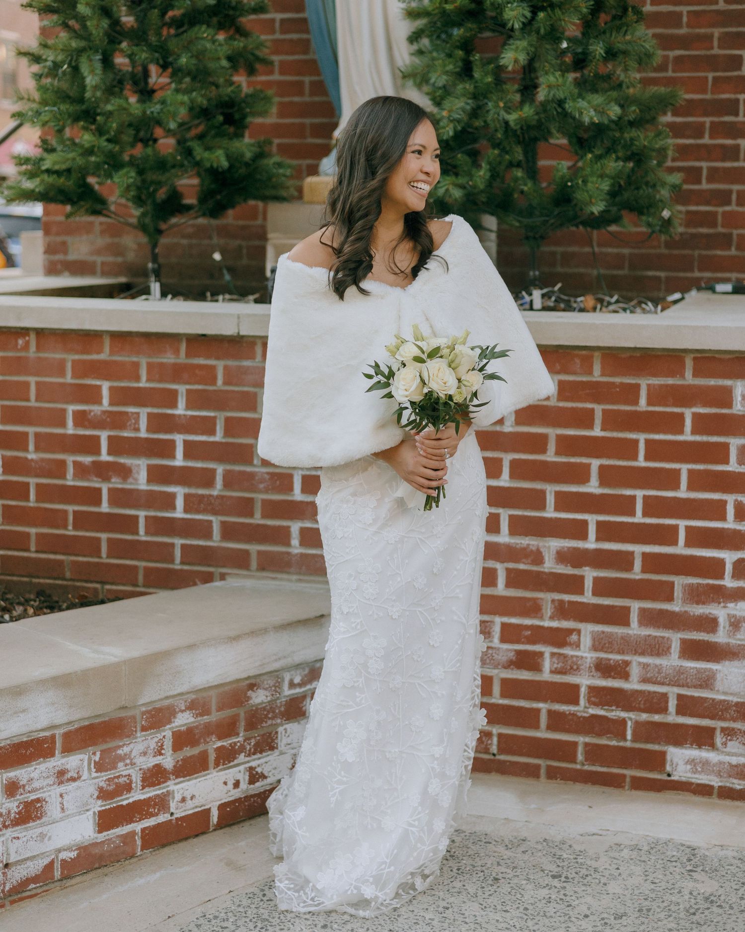 Bride in lace dress holding white bouquet against brick church wall with evergreen trees.