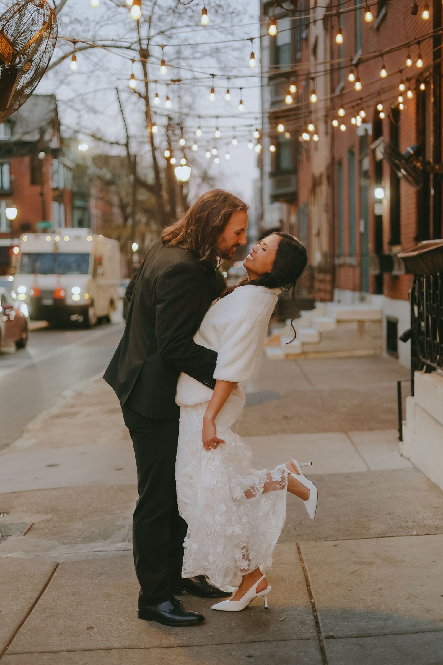 A playful wedding couple dances and twirls on an urban sidewalk decorated with string lights.