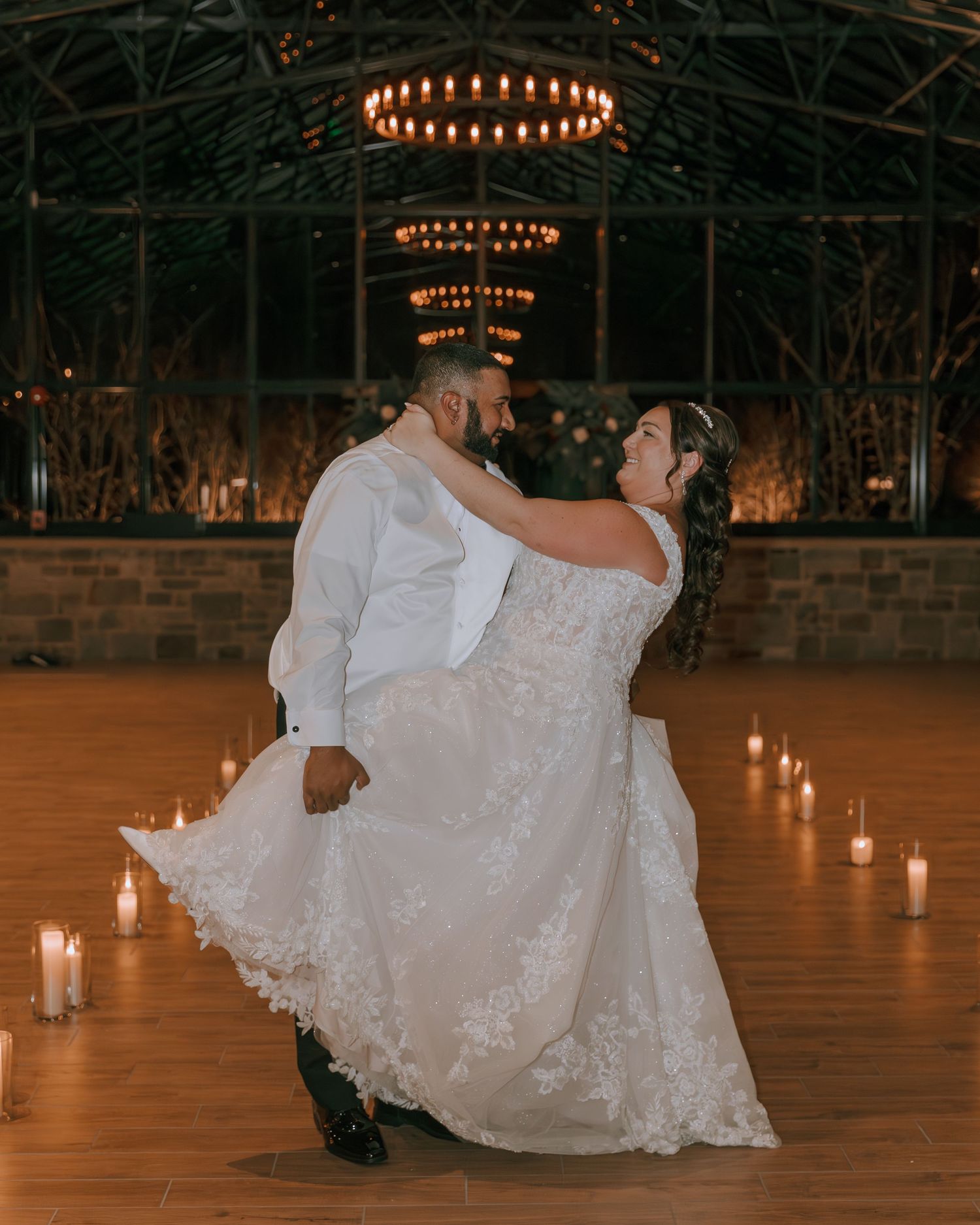 A couple dances romantically on a wooden floor under warm chandelier lighting in an elegant rustic venue.