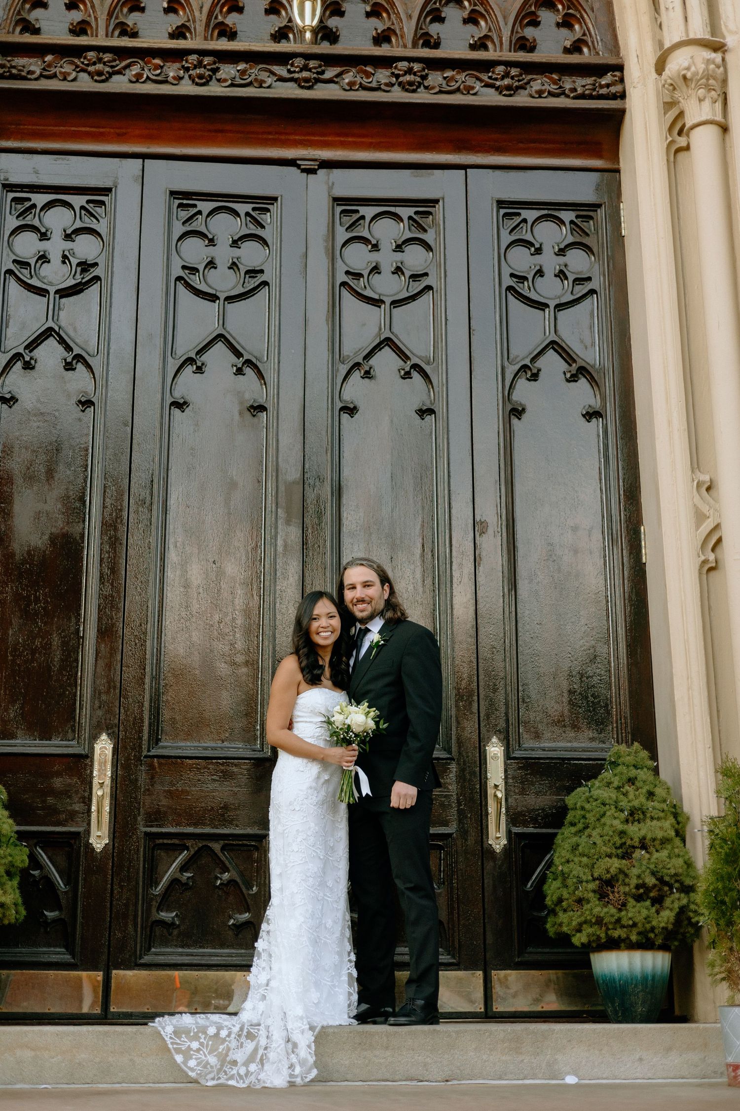 A couple in wedding attire poses in front of ornate Gothic-style doors with decorative metalwork.