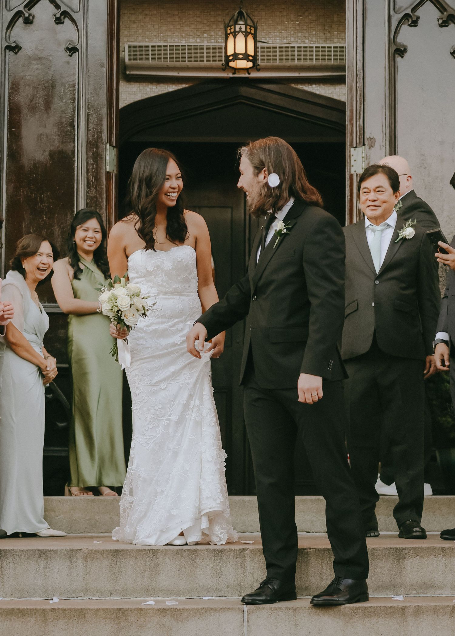 Wedding party members in formal attire gather on ornate stone steps during an outdoor celebration.