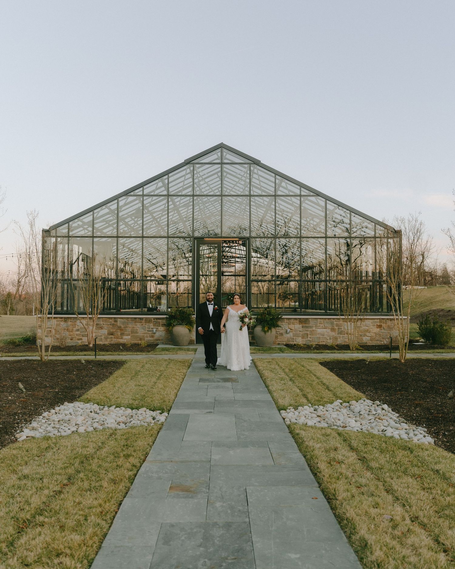 A wide shot of newlyweds standing before a grand glass greenhouse structure.