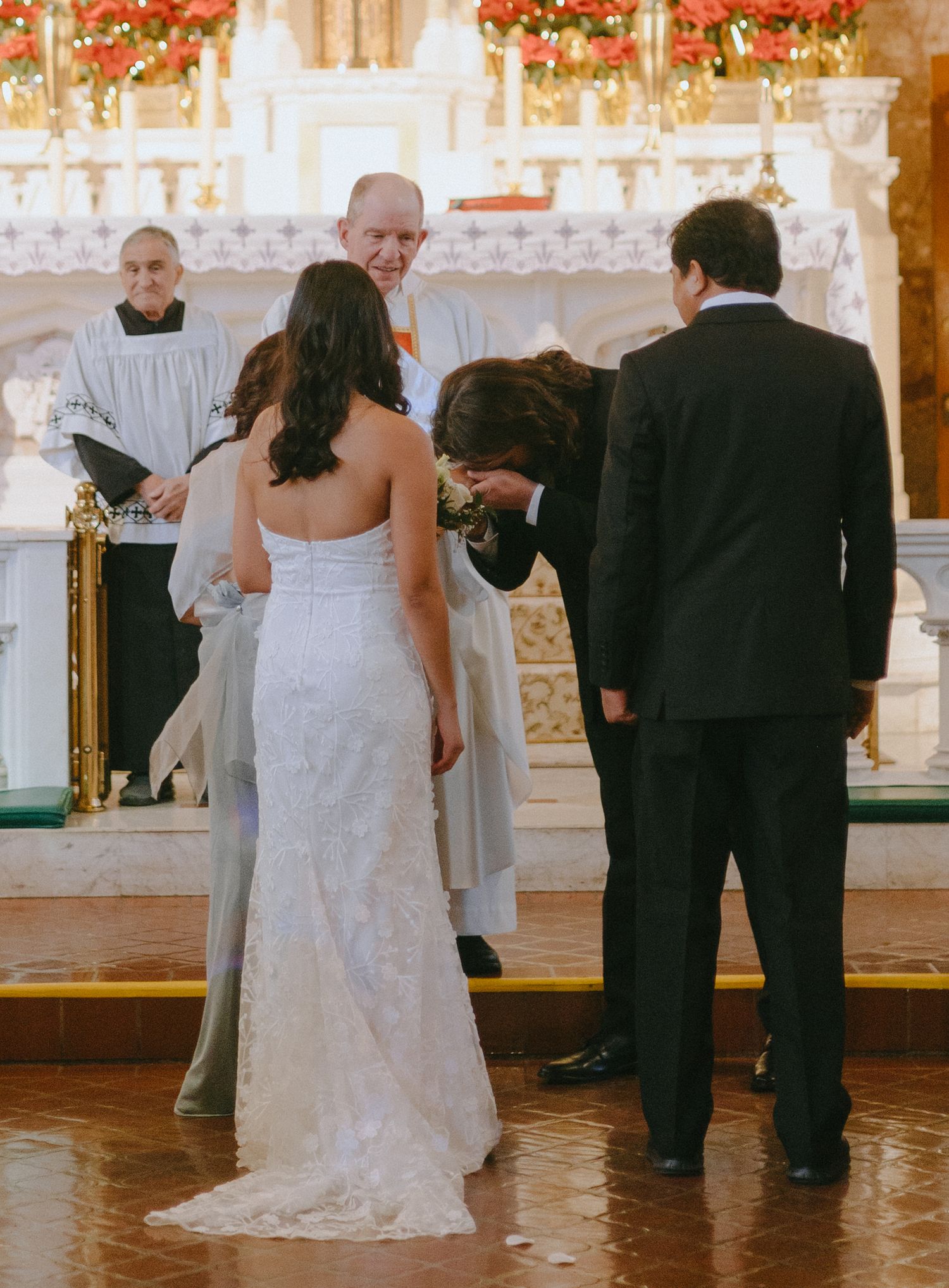 Wedding ceremony takes place in ornate church with red floral decorations and white columns.