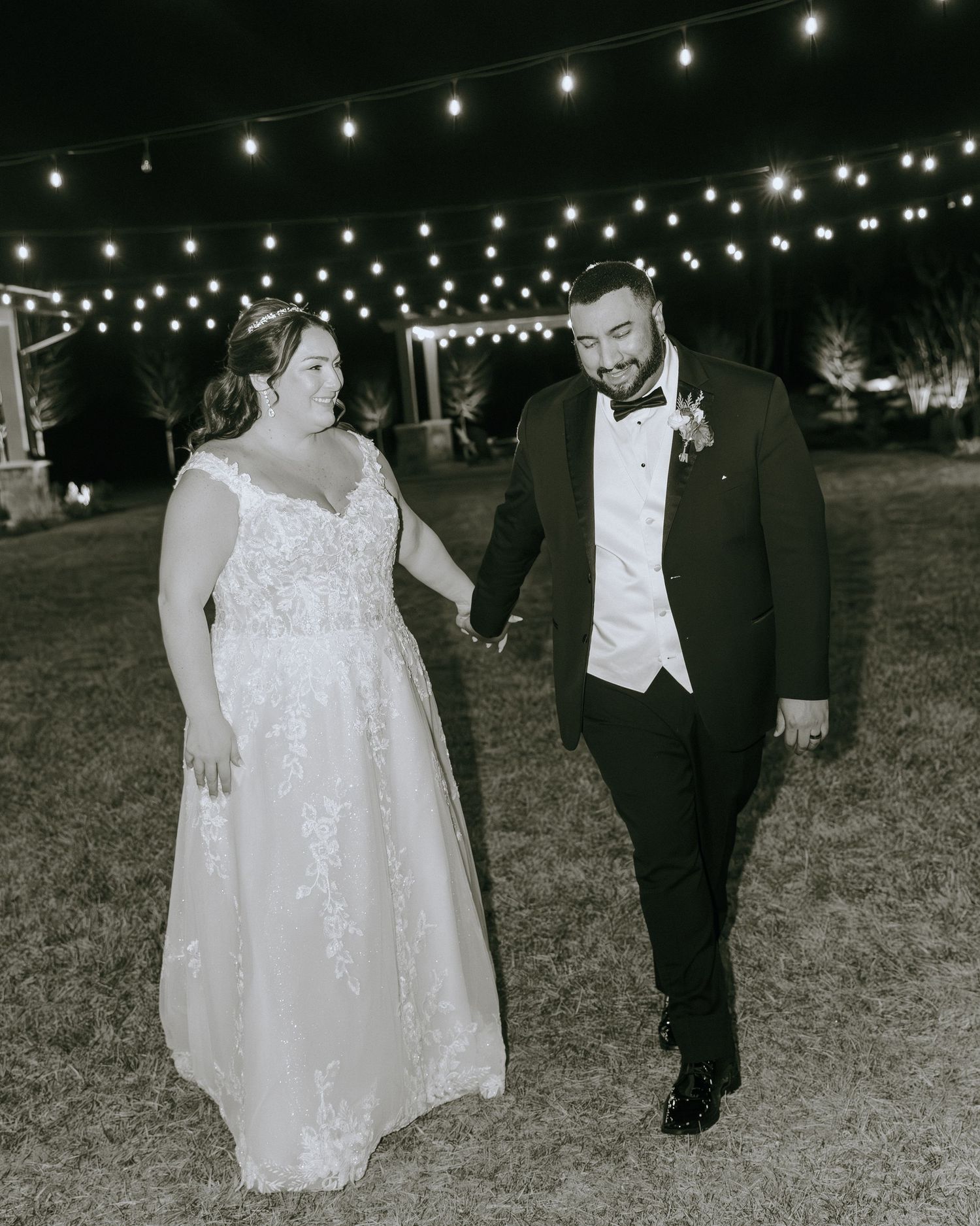 A black and white photo of newlyweds walking together under twinkling outdoor lights.