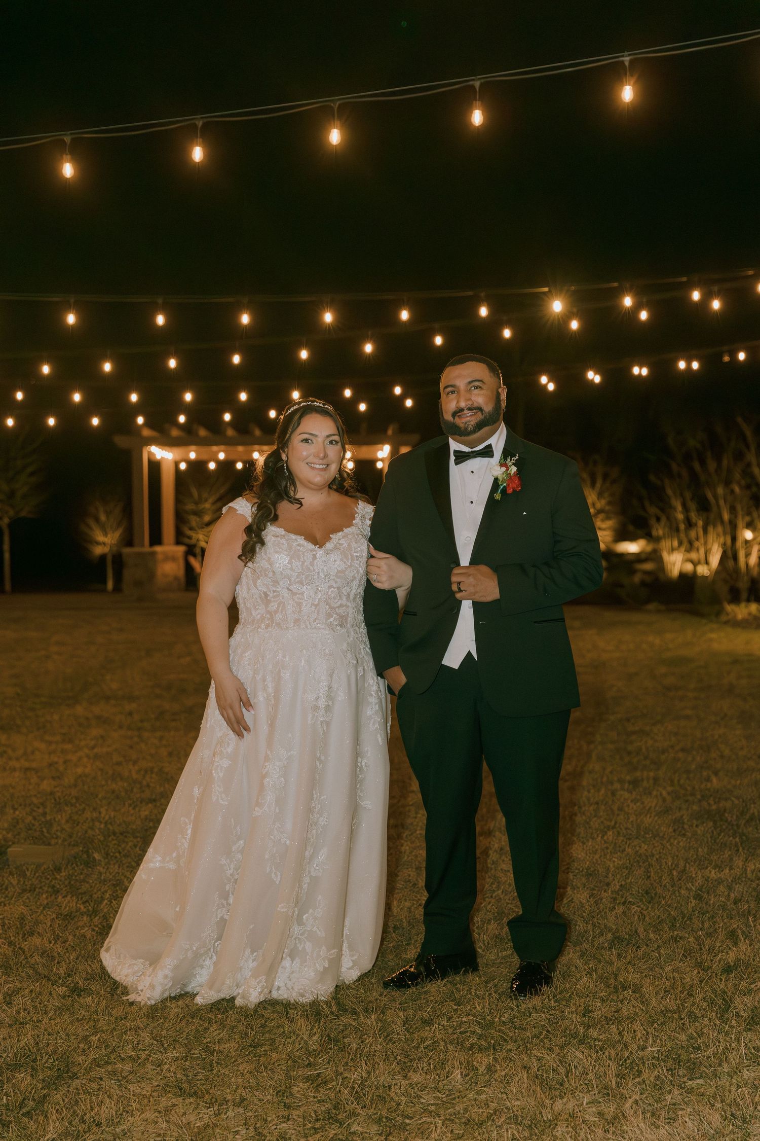 A wedding couple poses outdoors at night under romantic string lights.