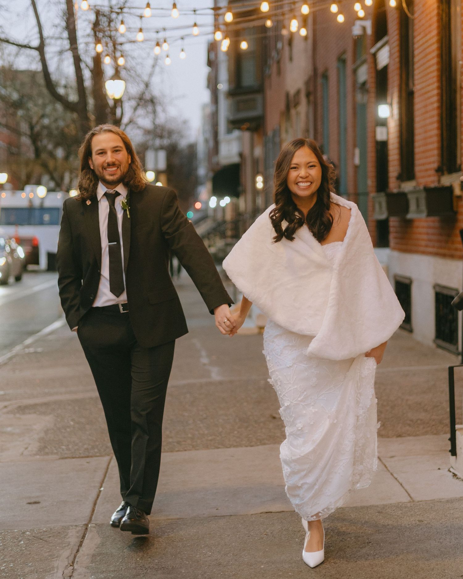 A couple in wedding attire walks down a city sidewalk with string lights overhead during a first look photo session.