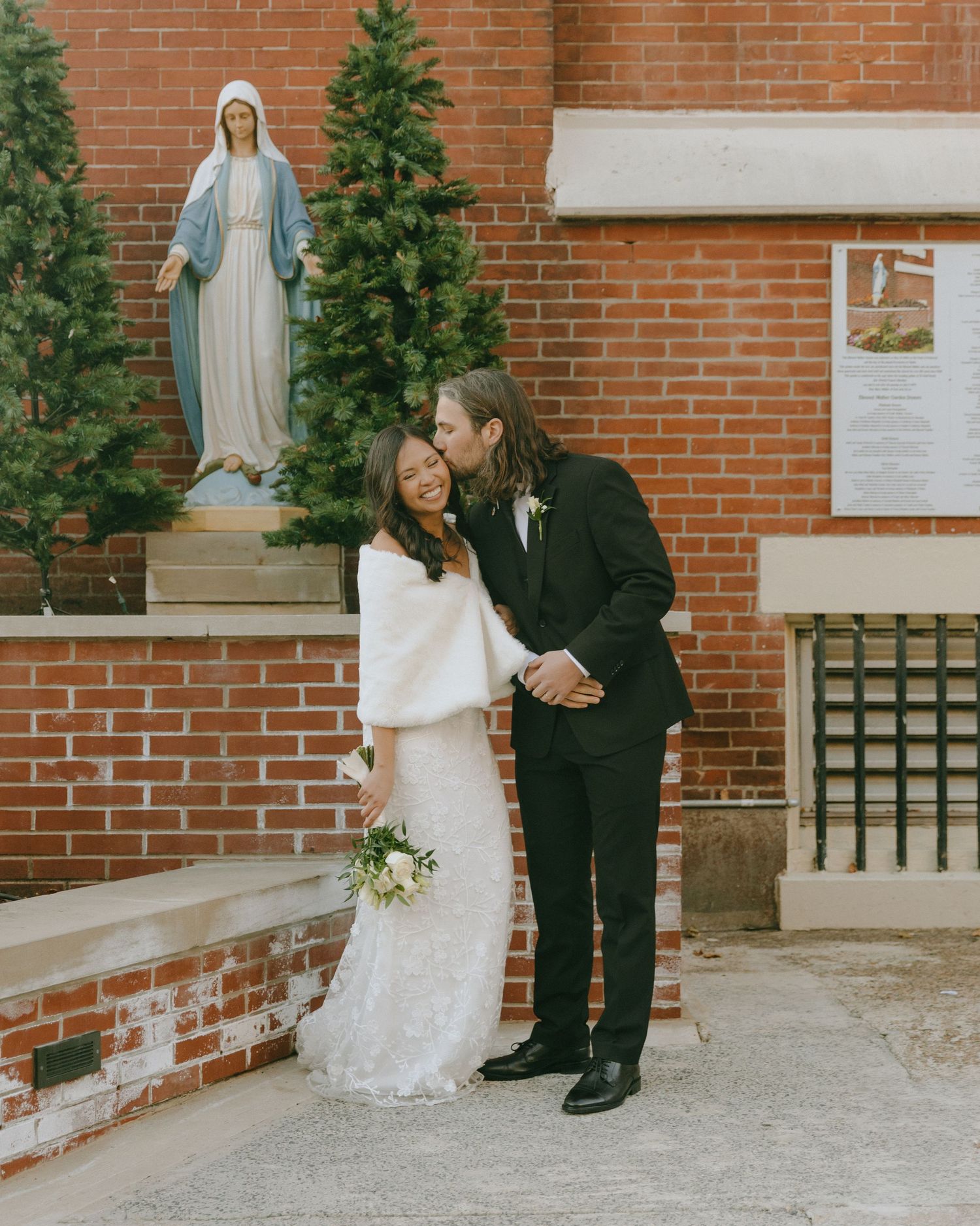 Couple sharing moment outside brick church with Christmas trees and statue of Mary.