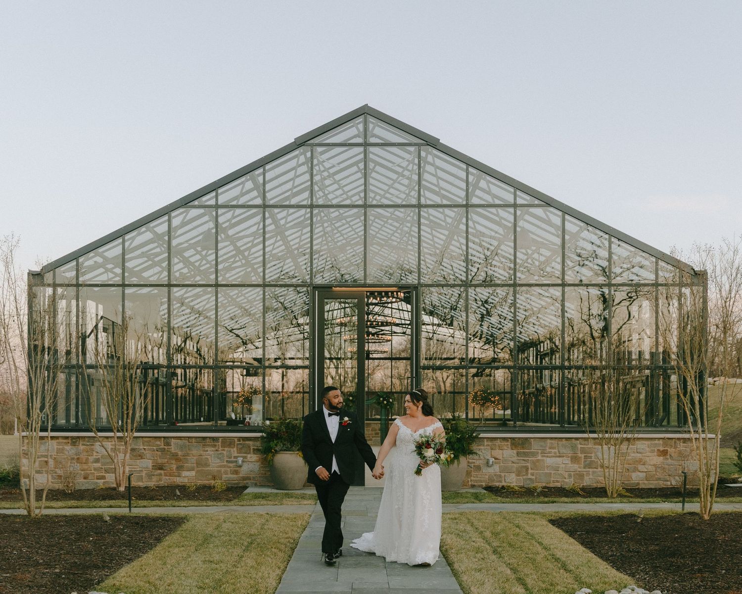 A couple poses in front of a modern glass greenhouse on their wedding day.