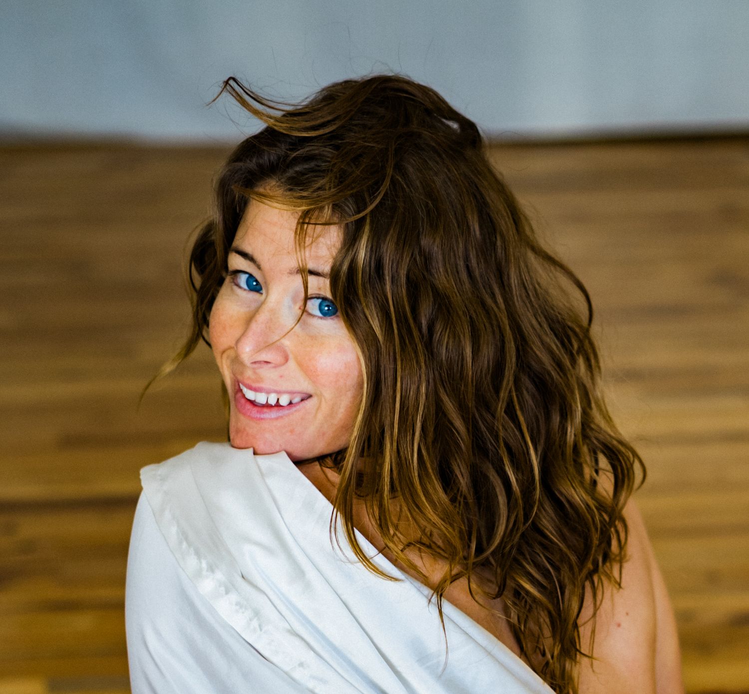 A person with windswept brown hair wearing a white top poses playfully against a wooden backdrop.