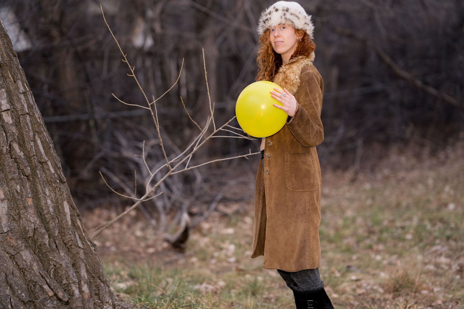 Person in vintage attire holds yellow balloon against bare winter trees.