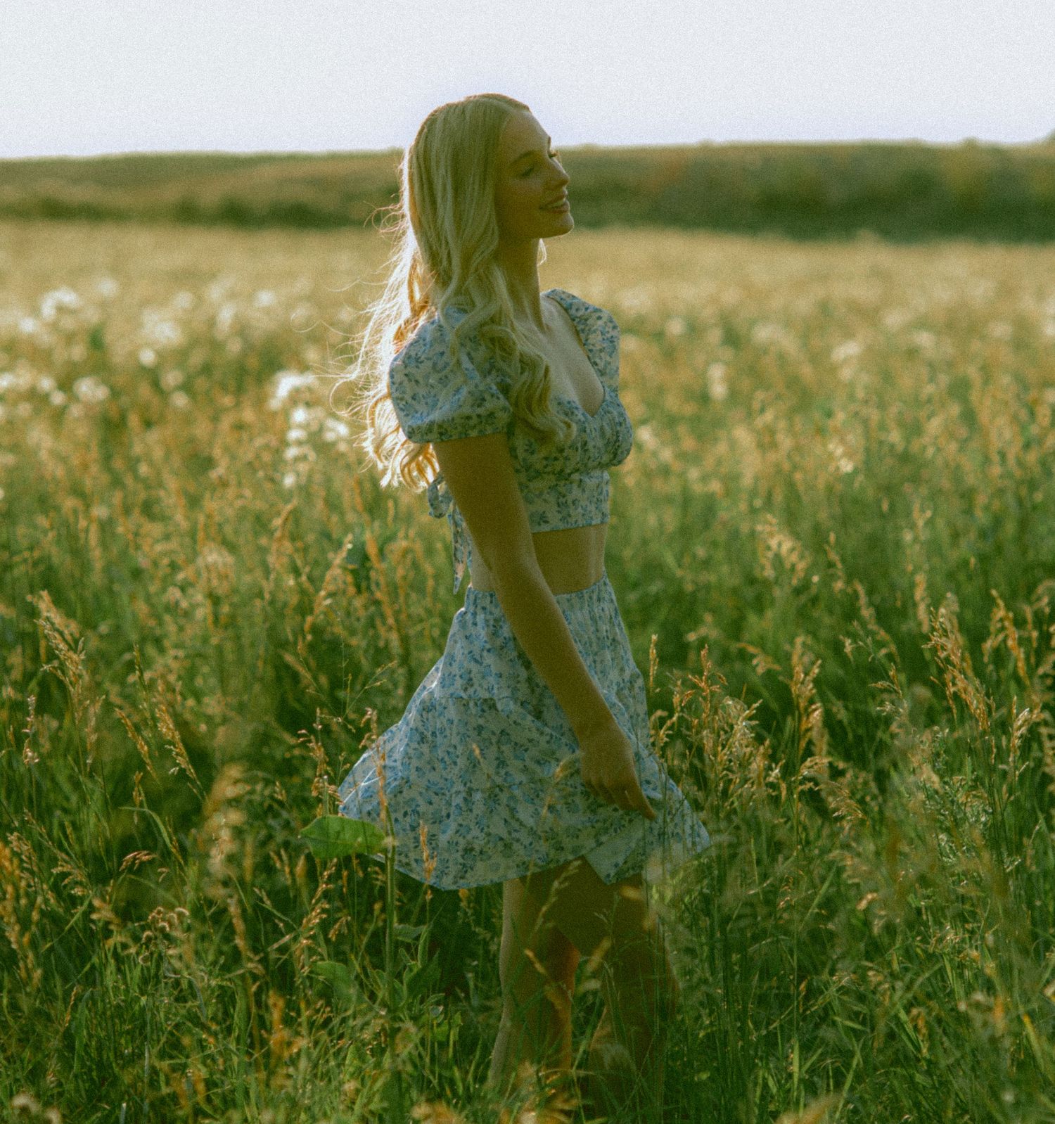 A person in a floral dress walks through a sunlit meadow of tall grass at golden hour for a senior session in Nebraska